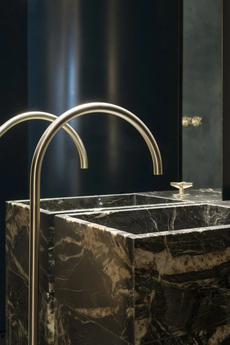 Close-up of a modern marble bathroom sink with a brushed gold faucet, dark blue wall in the background.