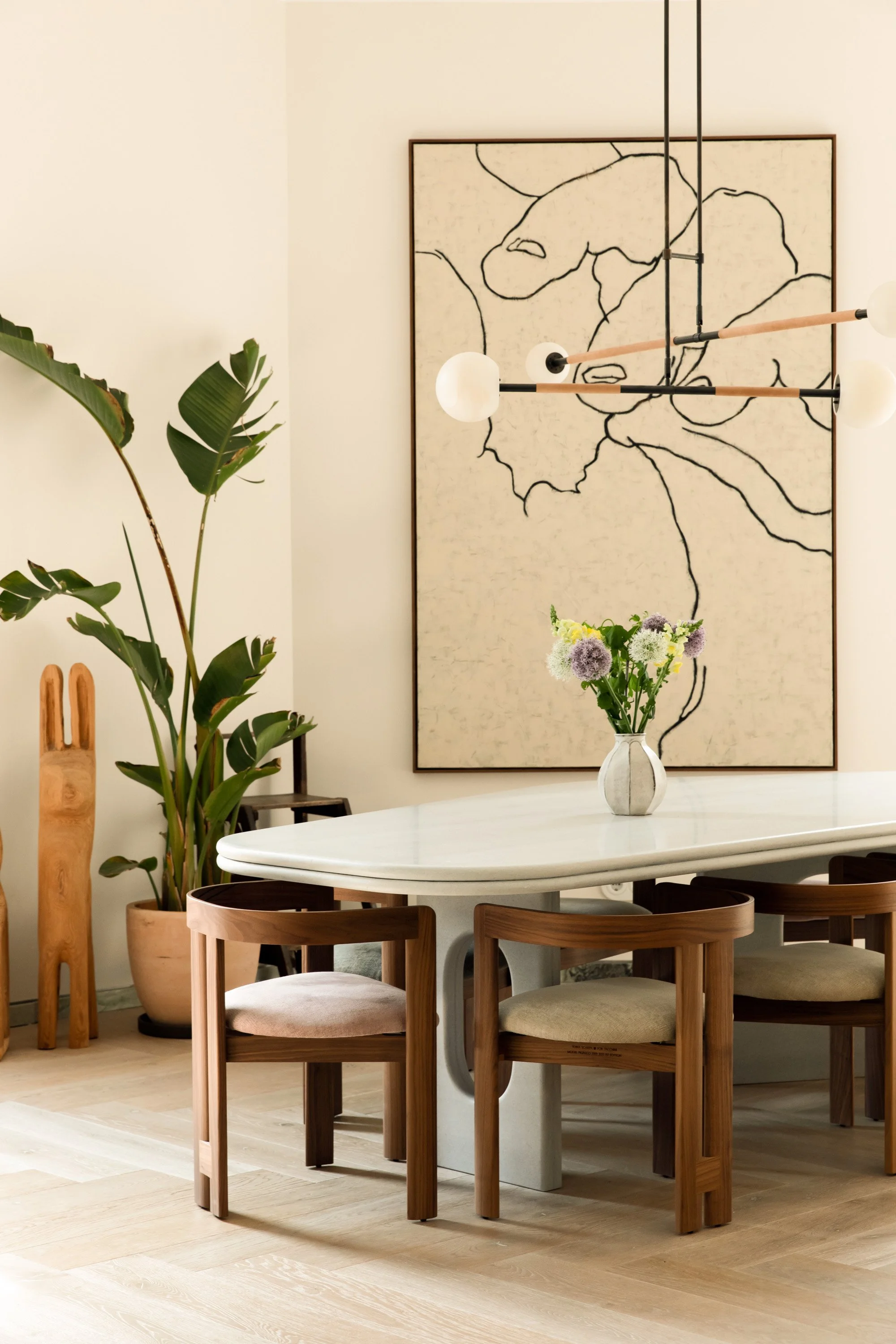Modern dining room with a white marble table, wooden chairs, a large potted plant, a vase with flowers, abstract wall art, and a contemporary chandelier.