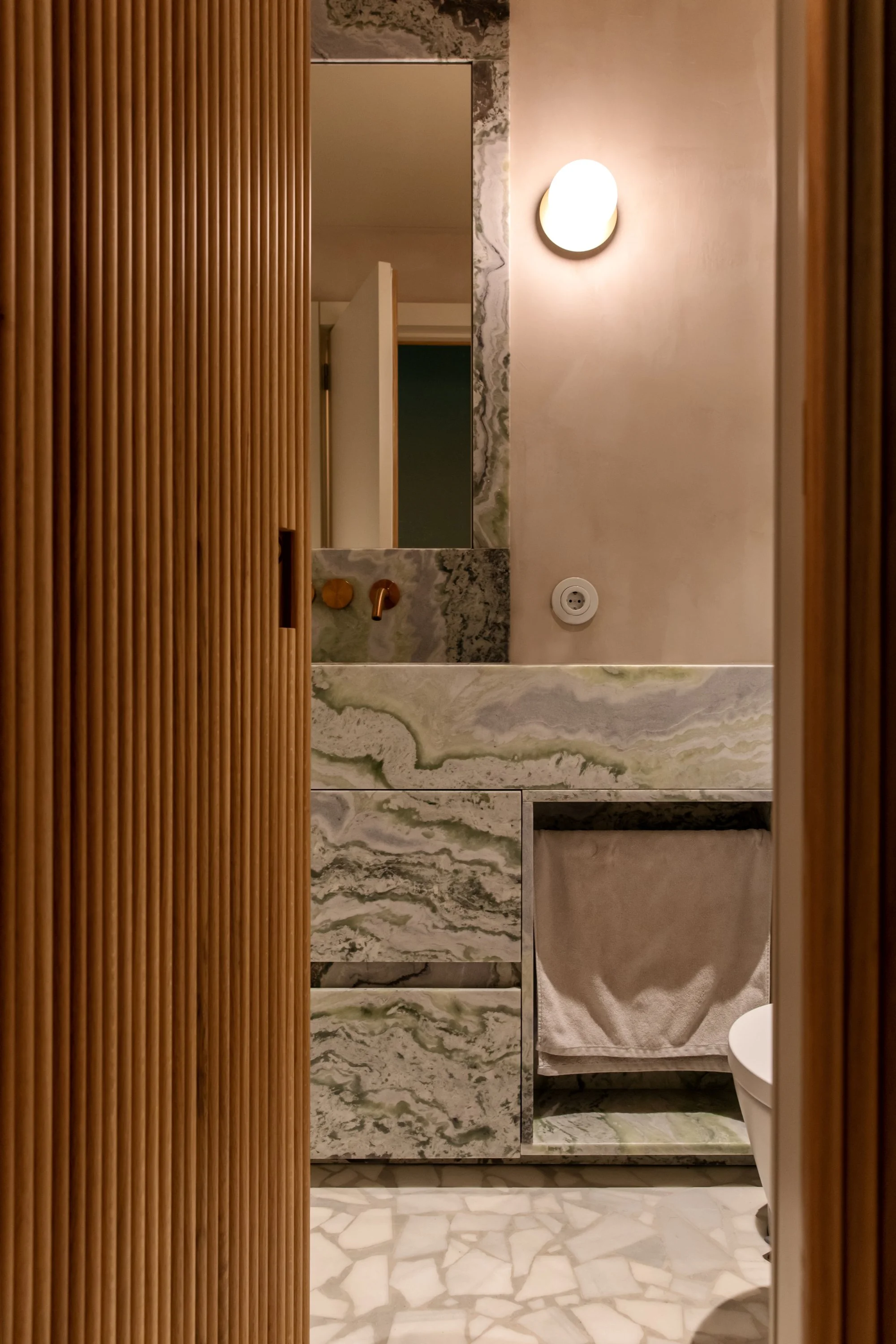 View of a modern bathroom with a marble sink, rectangular mirror, wall-mounted faucet, soft wall light, partial view of a toilet, and a sliding wooden door with vertical slats.