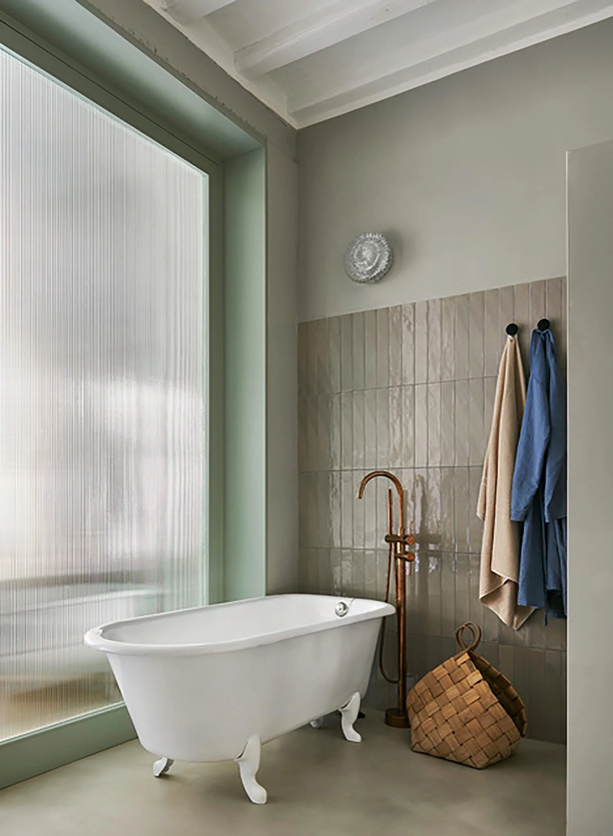 Bathroom with a vintage clawfoot bathtub next to frosted glass window, beige towel and blue towel hanging on hooks, wicker basket on the floor, copper floor lamp, and a decorative mirror above