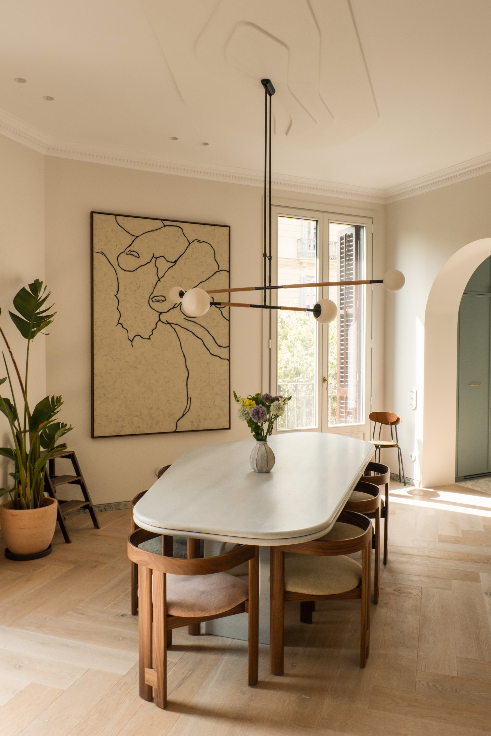 Bright dining room with a white oval table surrounded by wooden chairs, a vase of flowers, a large window letting in natural light, and a modern light fixture hanging from the ceiling.