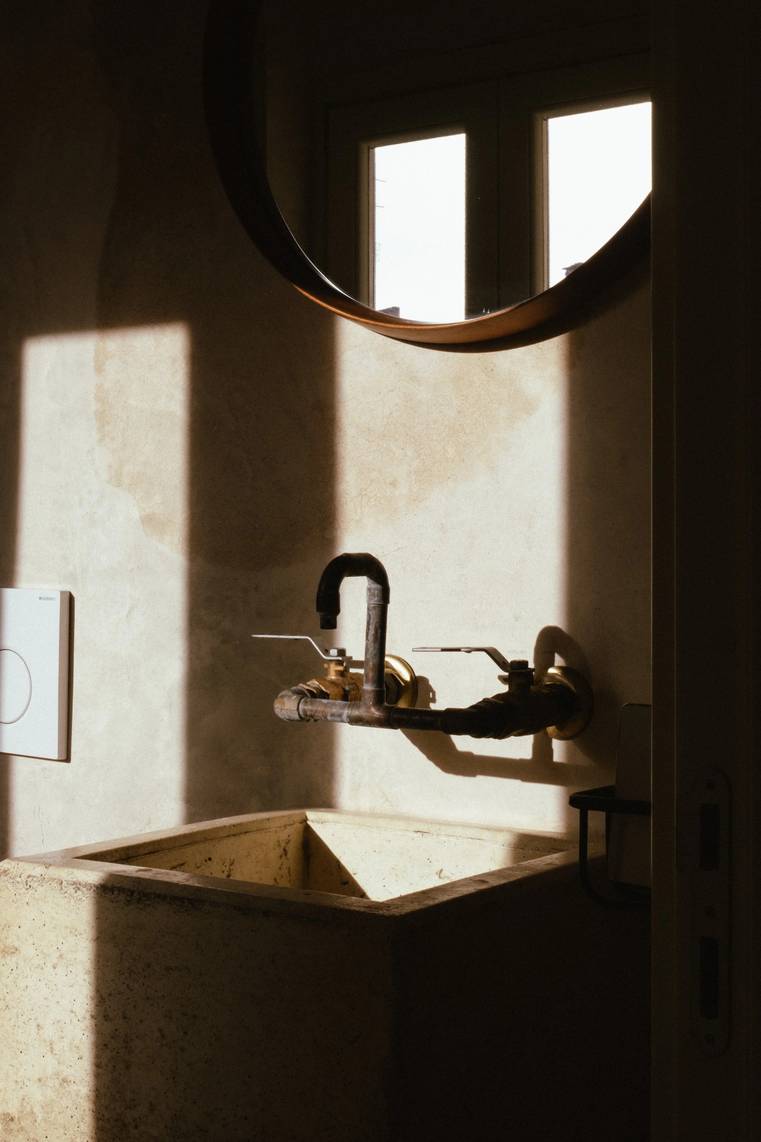 A rustic utility sink with a vintage faucet in a room illuminated by natural sunlight coming through a window, with a round mirror on the wall above.