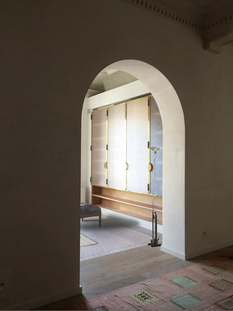 Interior view through an arched doorway showing a room with a large cabinet, a standing lamp, a bench, and a rug.