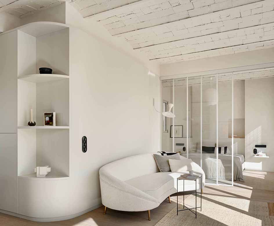 A modern, minimalist living space with white walls, a white curved sofa with pillows, a small side table, a white bookshelf with decorative items, and a glass partition separating the living area from a bedroom with a bed, lamps, and framed art.