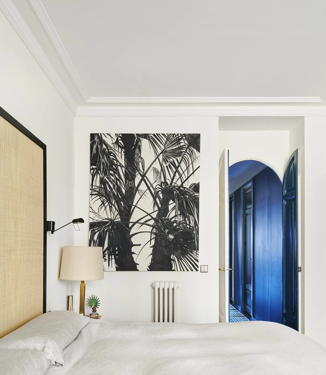A bedroom with a white bed, a beige headboard, a white wall with black and white tropical plant artwork, a small side table with a lamp and a green decorative object, and a doorway with blue doors leading to a hall.