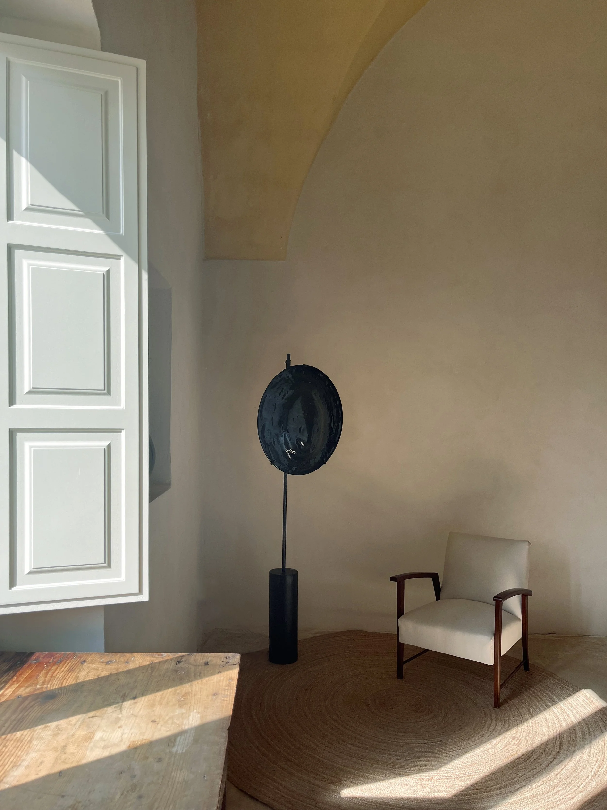 A corner of a room with a white chair, a black standing lamp, a circular beige rug, a wooden table on the left, and an open white cabinet door on the left side of the image.