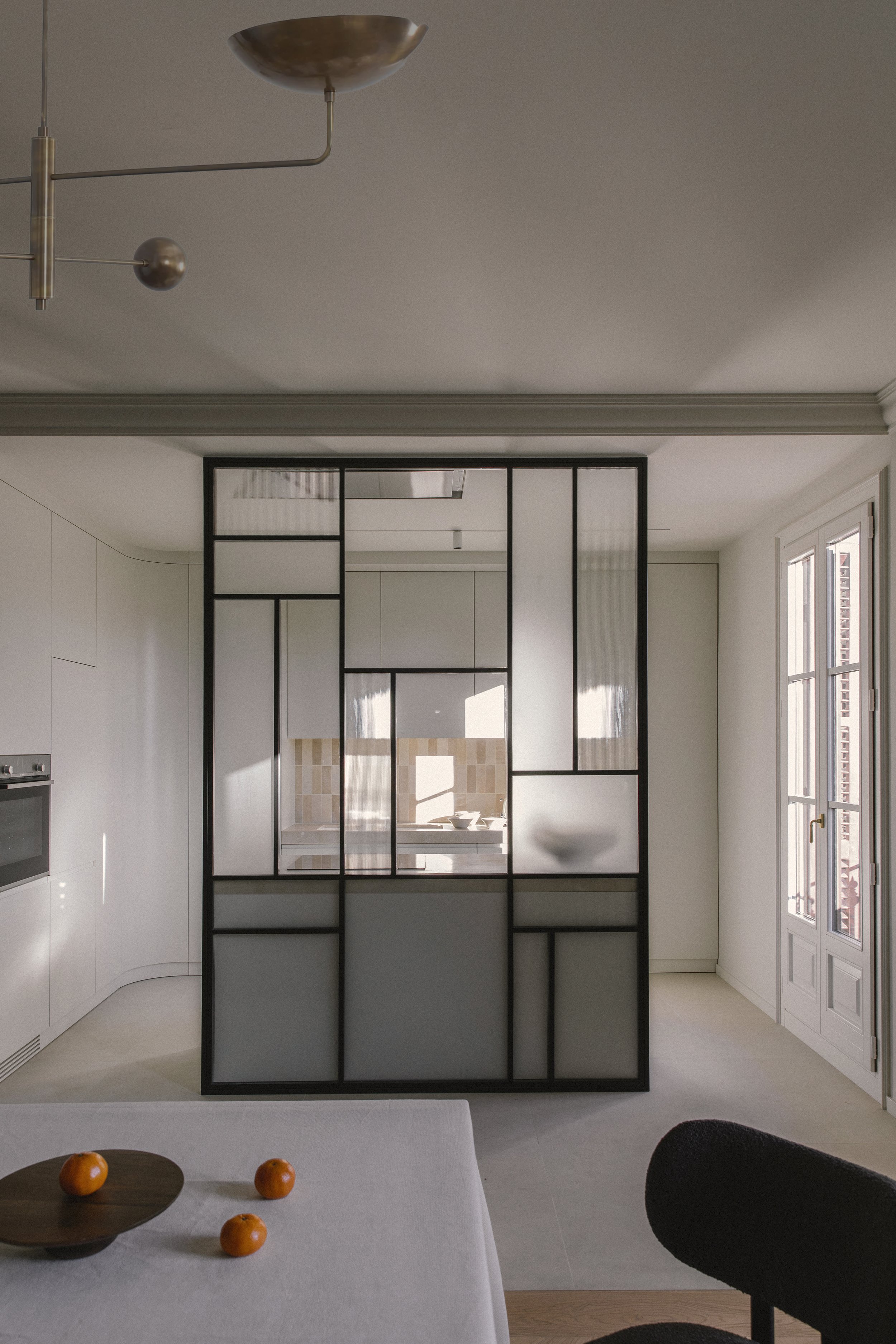 Interior view of a modern, minimalist kitchen and dining area with a decorative black-framed glass partition, dining table with three oranges and a wooden bowl, and a window letting in natural light.