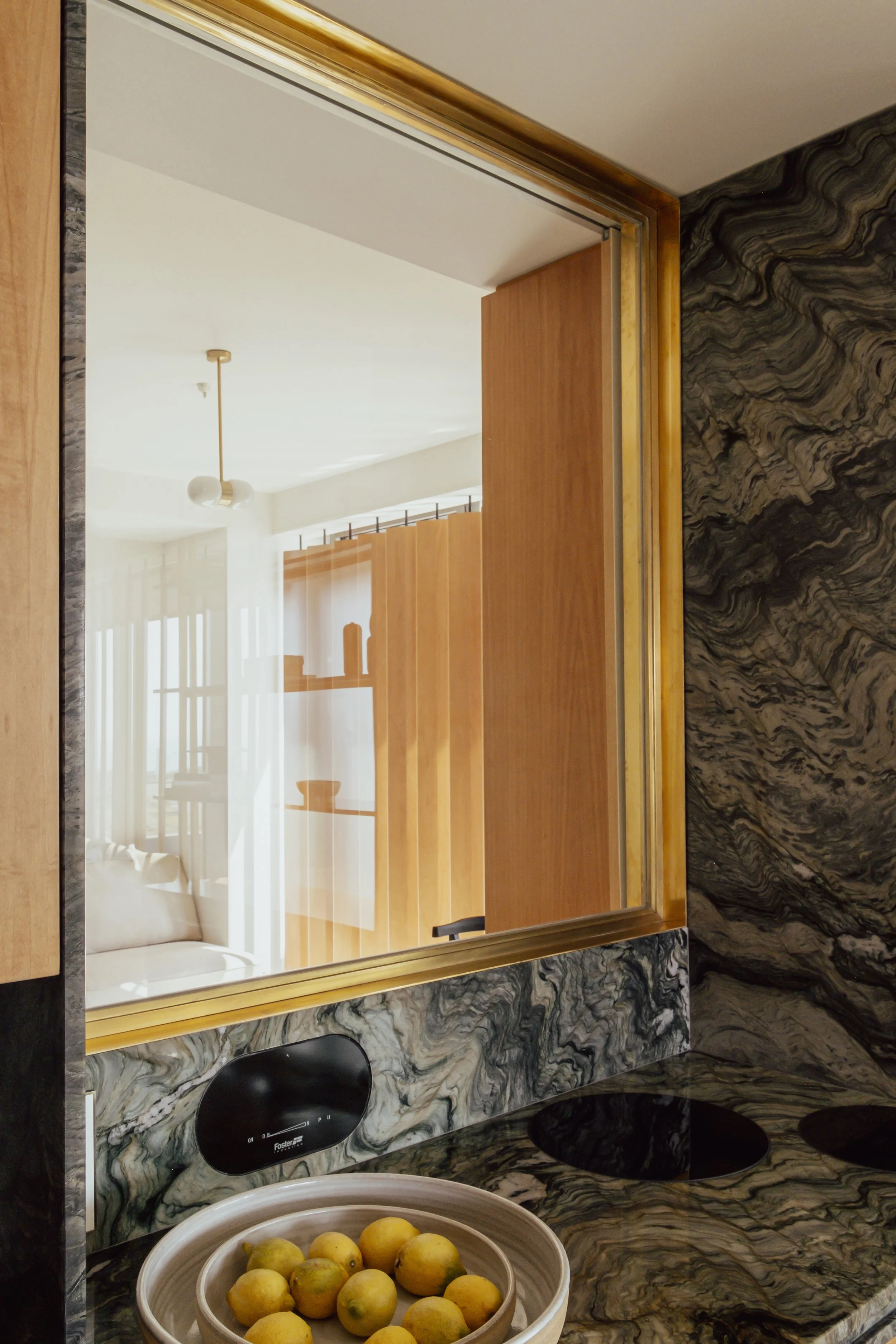 Close-up of a modern kitchen with a marble countertop and black induction cooktop, a bowl of lemons, and a large mirror reflecting a living area with wooden cabinets, a pendant light, and a window with vertical blinds.