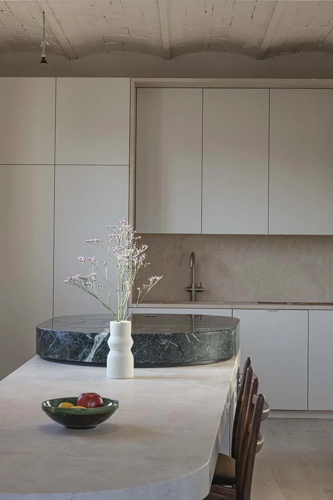 Minimalist kitchen with white cabinetry, beige backsplash, and a marble island with a vase of flowers and a bowl of fruit on top.