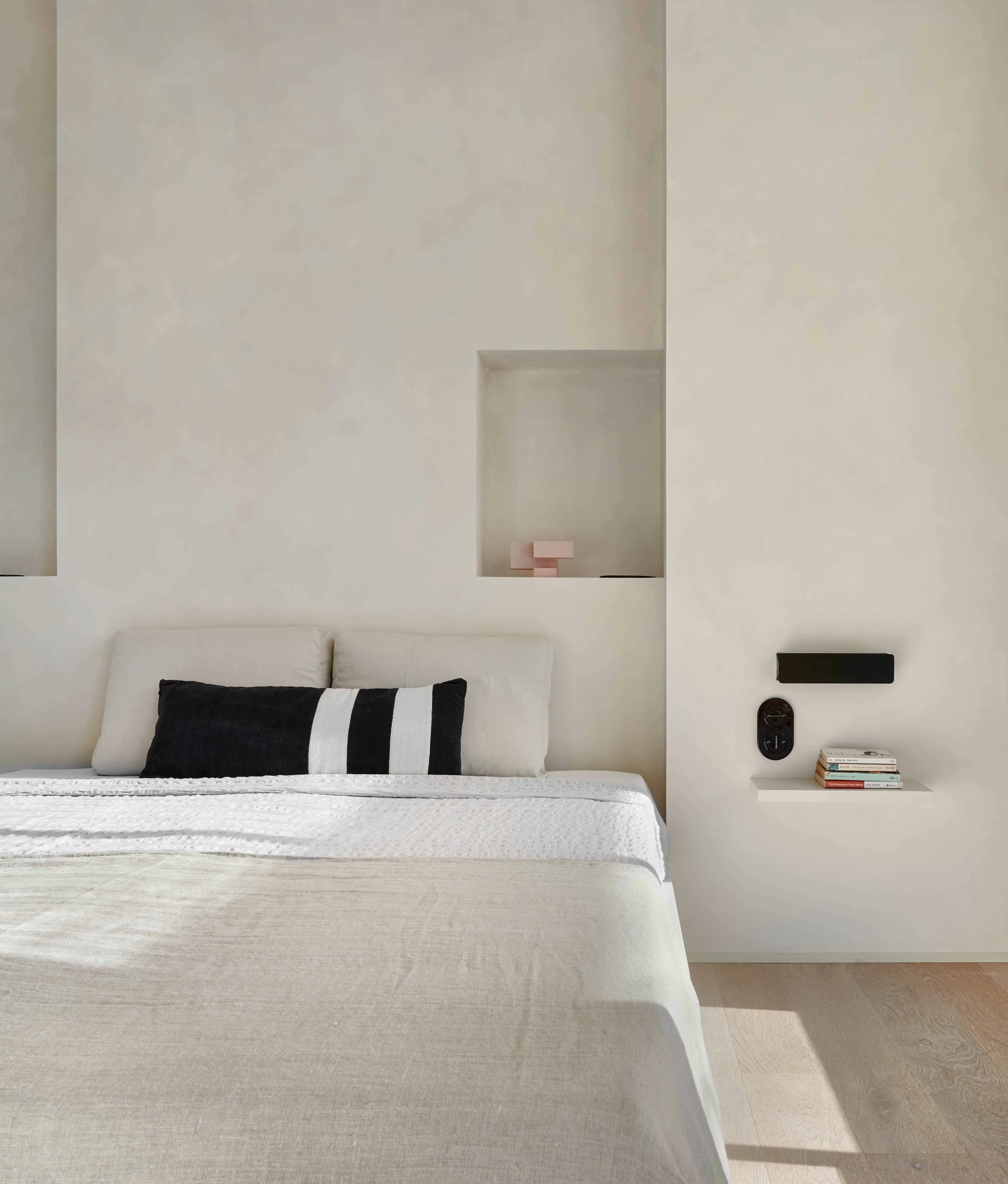 A minimalist bedroom with a white bed, a black and white striped pillow, a built-in shelf with pink objects, and a small white side table with stacked books and electronic devices.