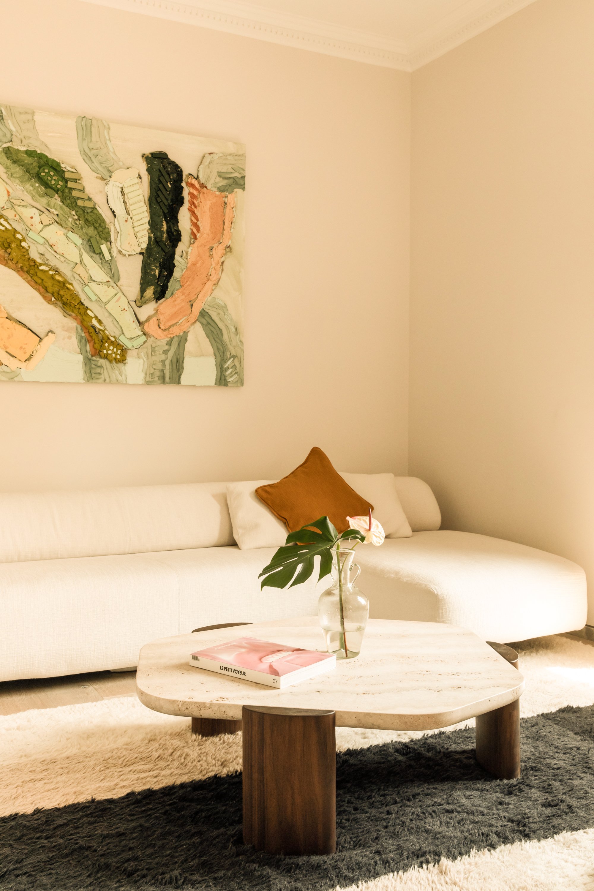 A living room featuring a white sofa with a brown accent pillow, a marble coffee table with a magazine and a glass vase containing large green leaves and a white flower, an abstract wall art piece, and a beige and black rug.