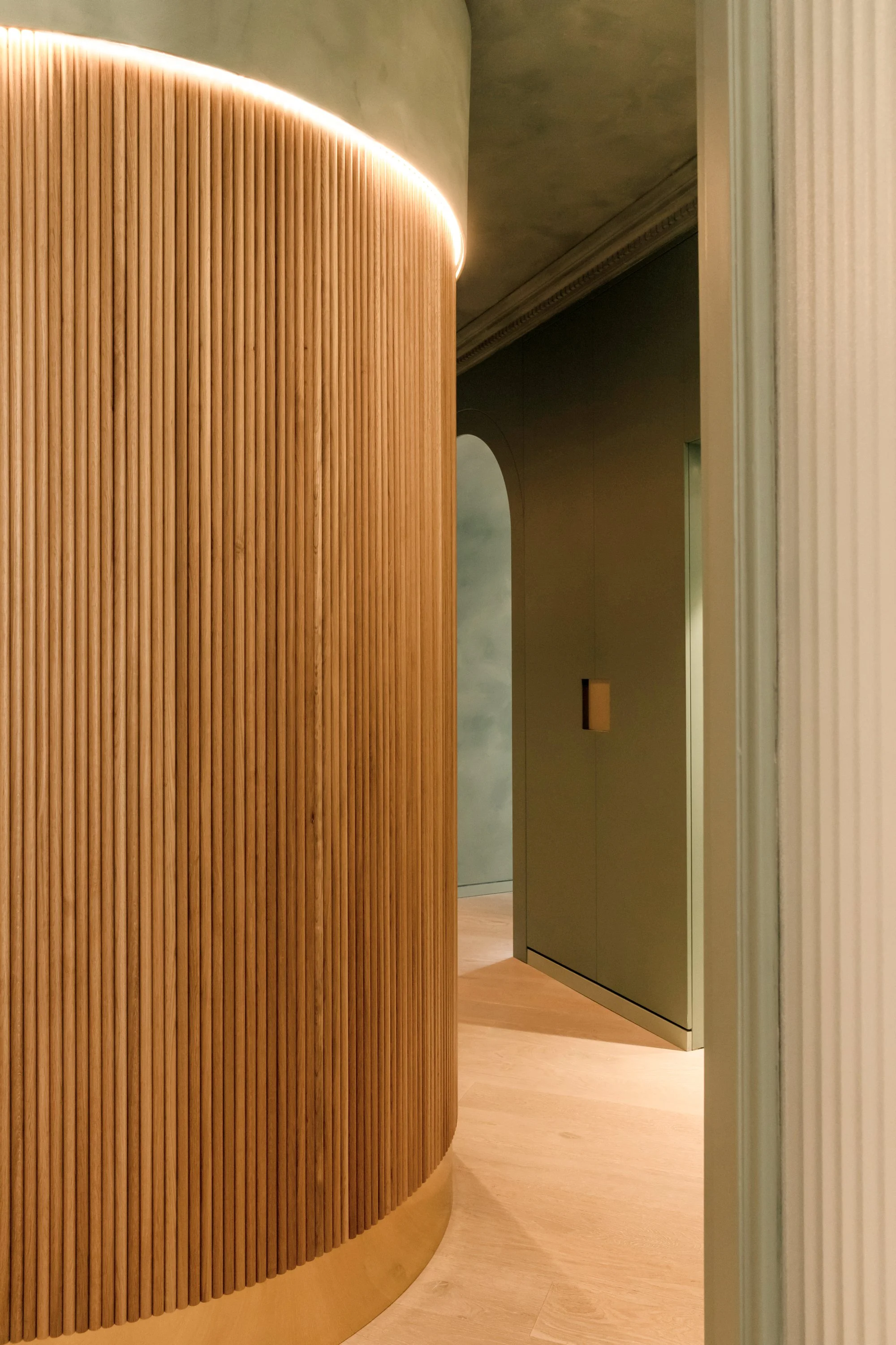 Interior view of a modern space with curved wooden slat wall and minimal decor, with a partial view of a doorway and a muted color palette.