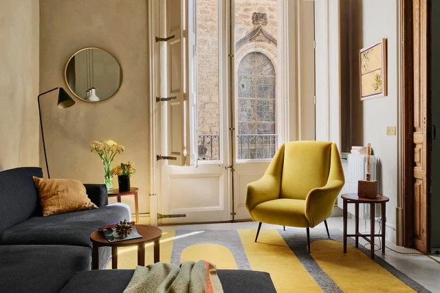 A cozy living room with a black sofa, yellow armchair, wooden side tables, and a large window with a view of a historic stone building outside.