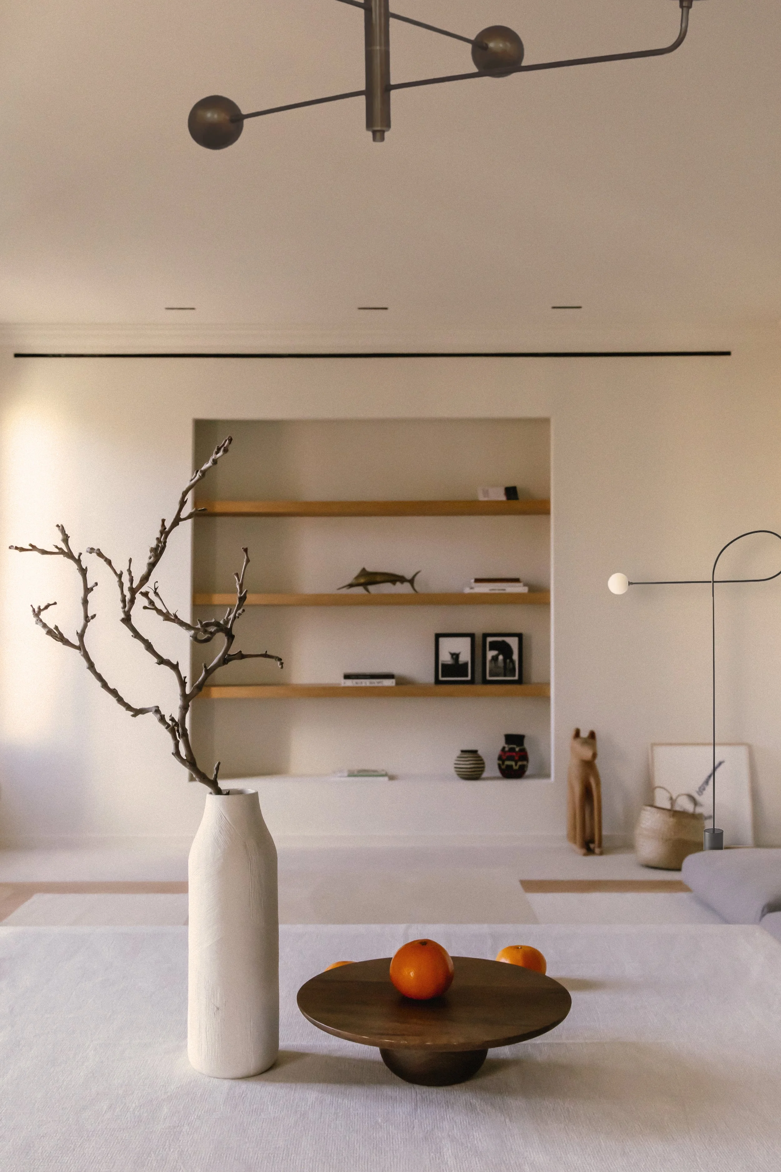 Minimalist living room with a white vase holding a leafless branch, a small wooden bowl with three oranges on a table, built-in wooden shelves with decorative items and framed pictures, and modern lighting fixtures.