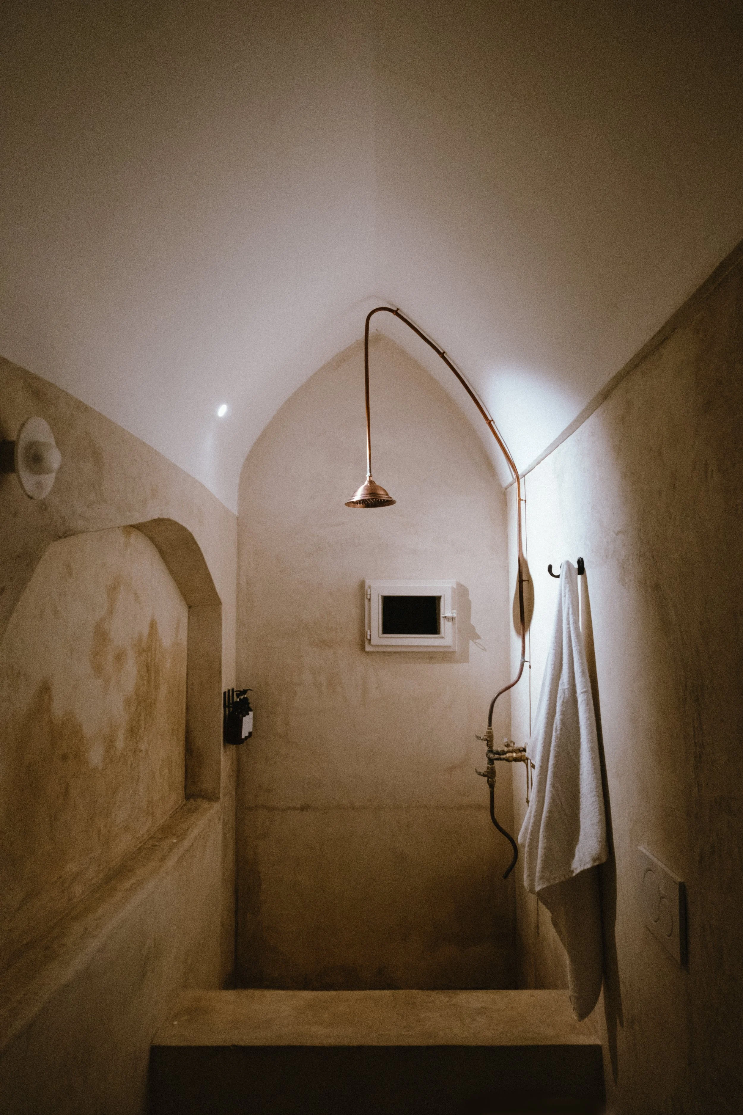 An unfinished shower area with beige plaster walls, a small window, a flex shower head hanging from the ceiling, and a white towel hanging on a hook.