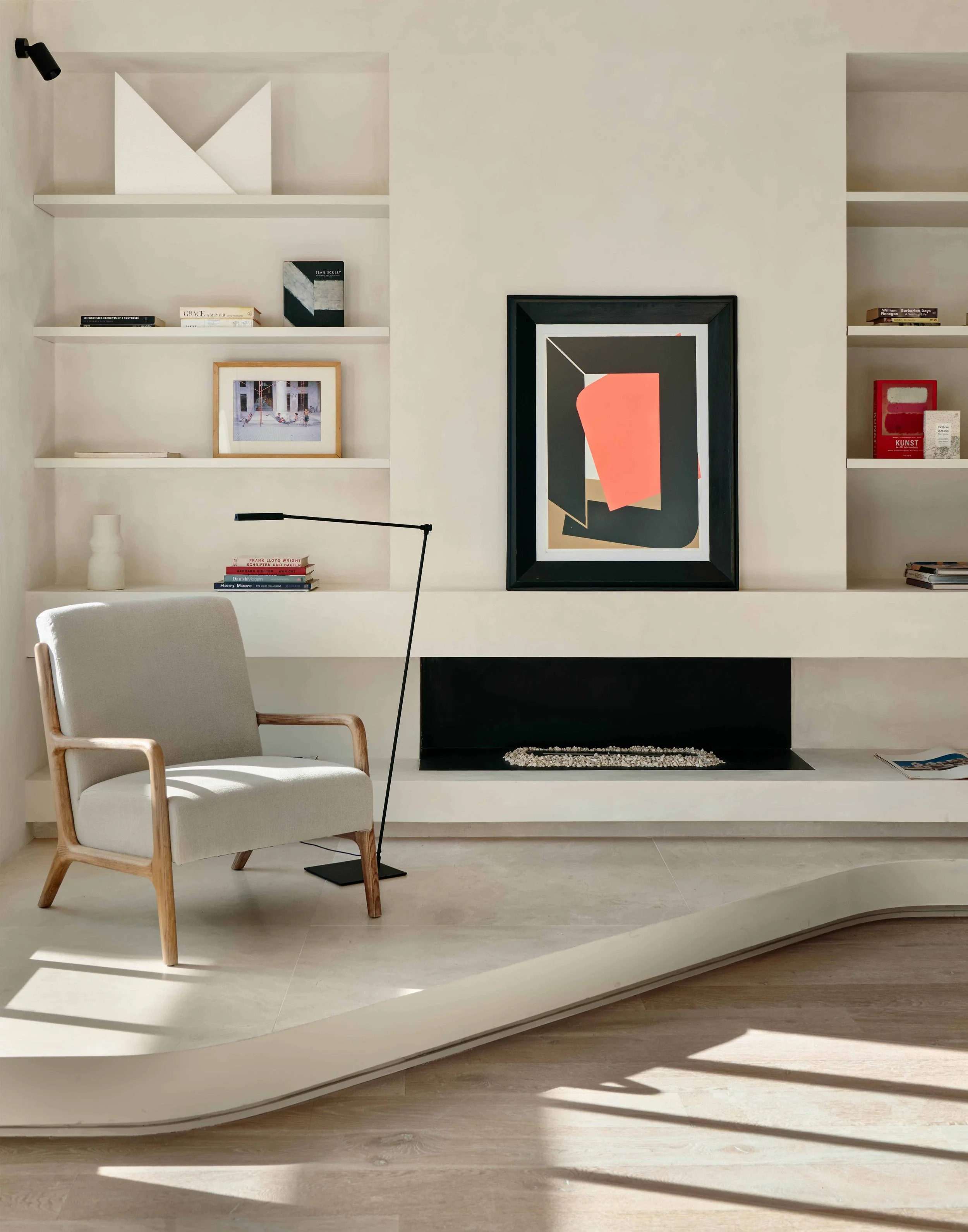 Modern living room with white built-in shelves, framed artwork, a white armchair with a wooden frame, a black floor lamp, and a minimalist black fireplace with decorative stones, all illuminated by natural light.