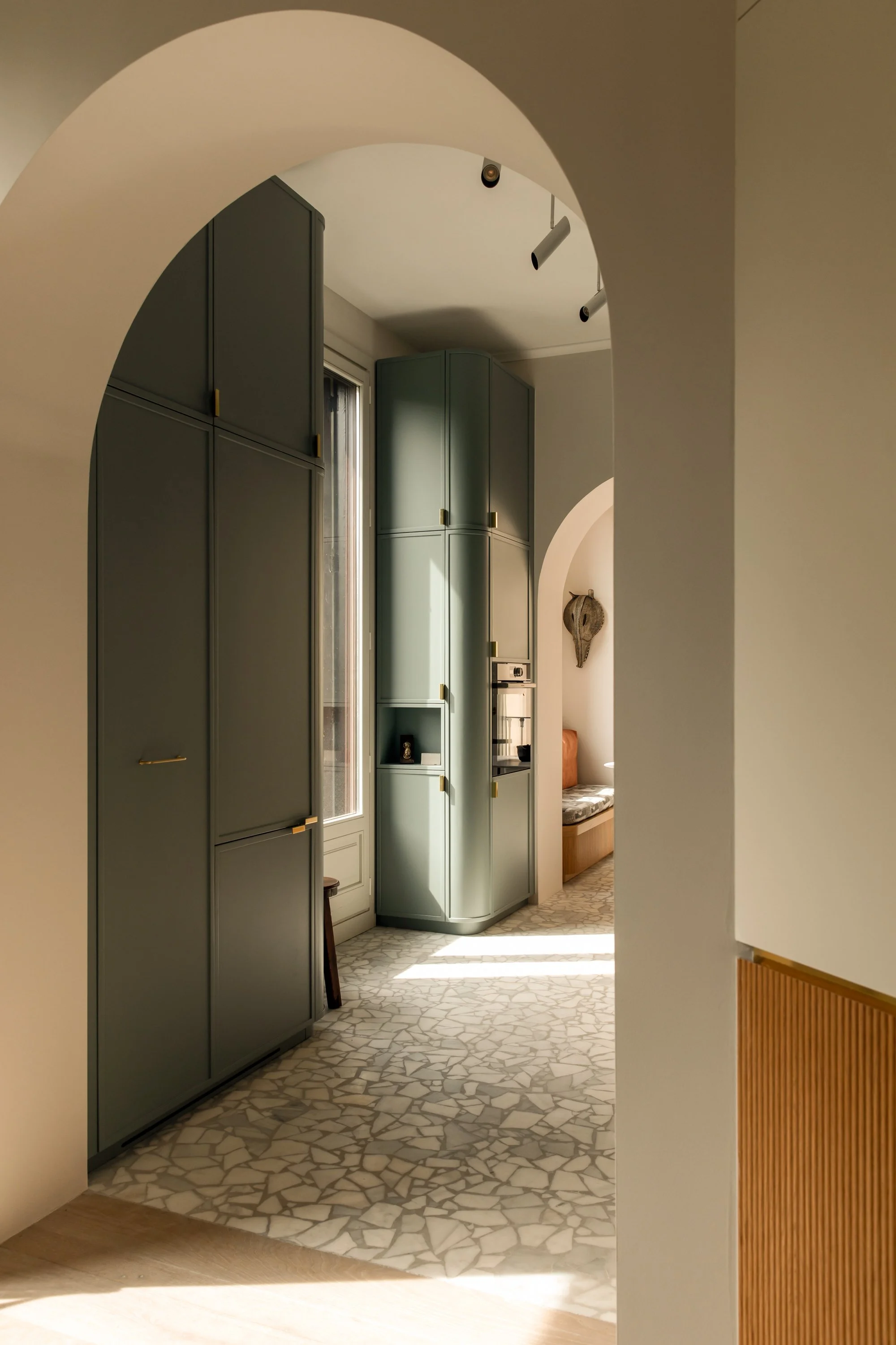 A shot of a hallway with arched doorways, showing built-in storage cabinets in mint green, a window with sunlight streaming in, and a sitting nook with a chair and decorative wall art.