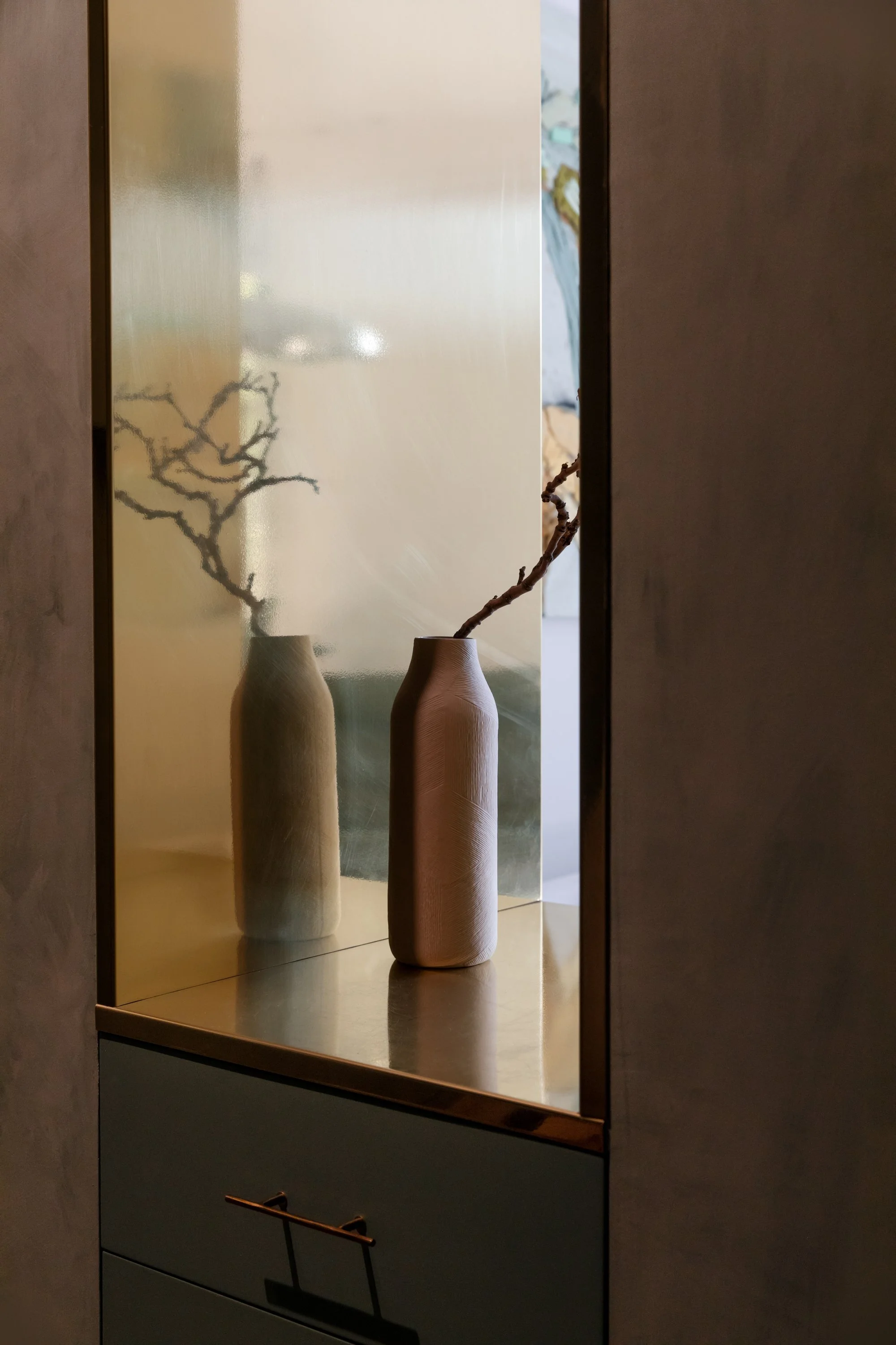 Two ceramic vases with branches, one missing its top, displayed on a wooden surface with a reflective golden back in a modern decor setting.