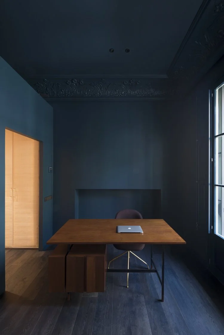 Interior of a minimalist office with dark blue walls, wooden desk with a laptop, and a black chair, lit by natural light from large window.