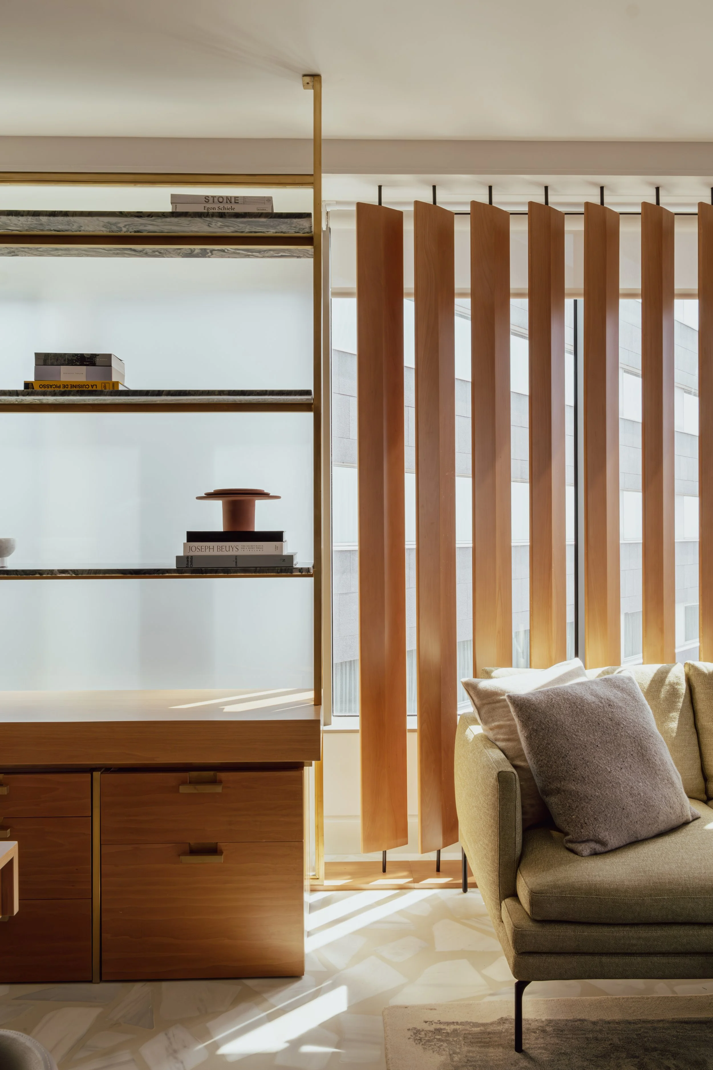 Interior of a modern living space with wooden shelves, a beige sofa with cushions, vertical wooden slats, and large windows bringing in natural light.