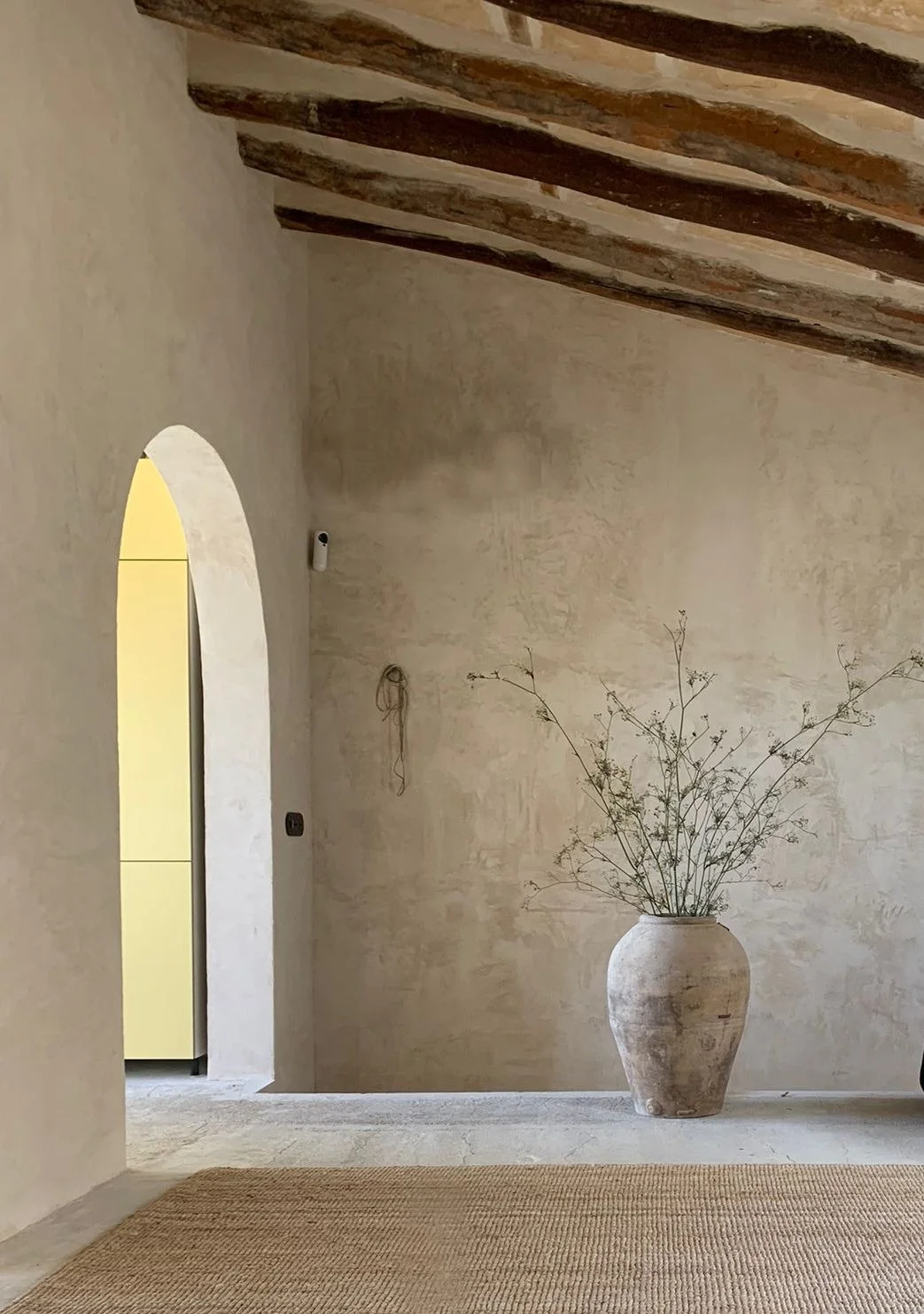Interior scene featuring a beige textured wall, wooden ceiling beams, a large ceramic vase with dried branches, an arched doorway, and part of a yellow cabinet visible through the opening.