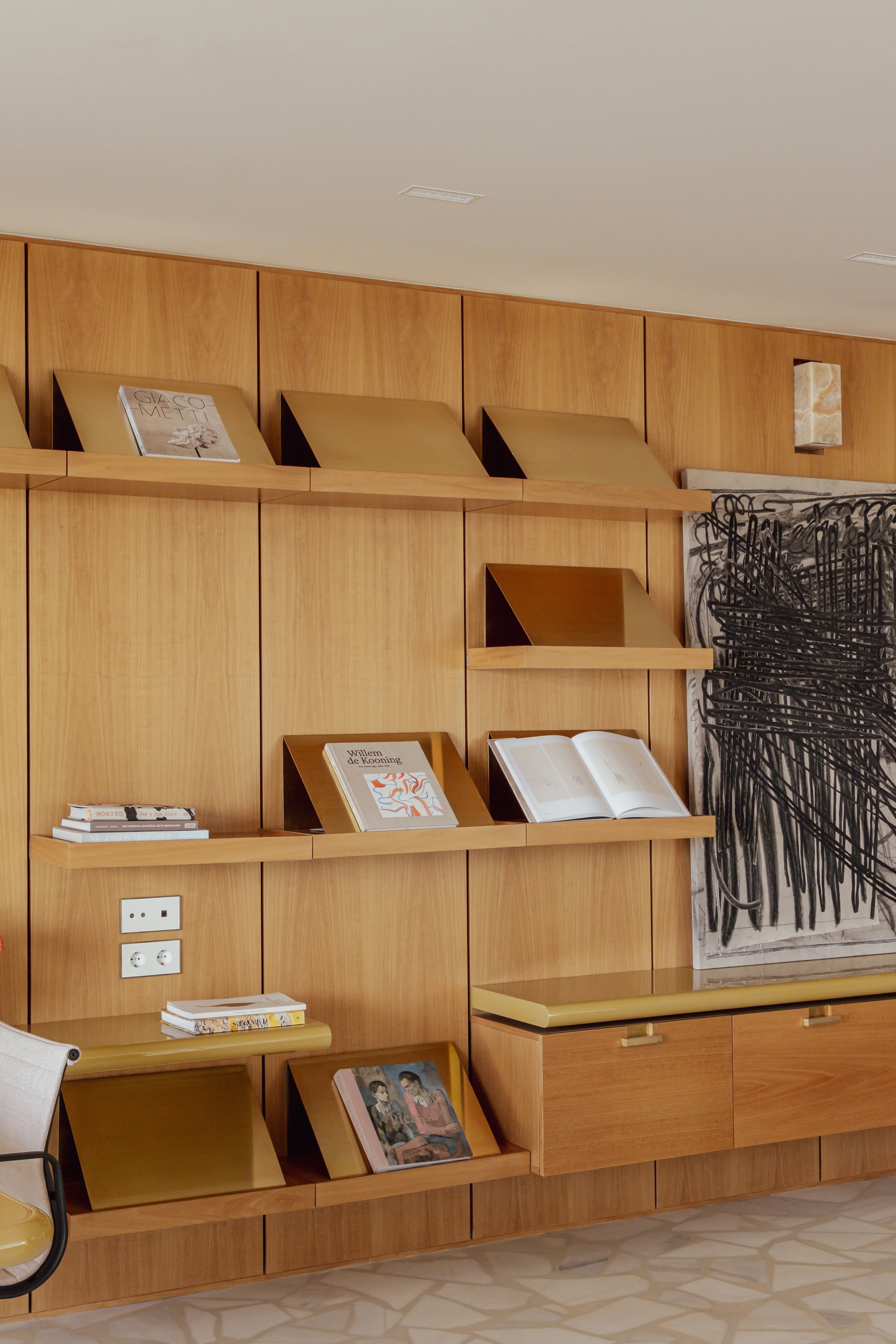 Wooden bookshelf with angled shelves holding books and magazines, and an abstract black and white artwork to the right.
