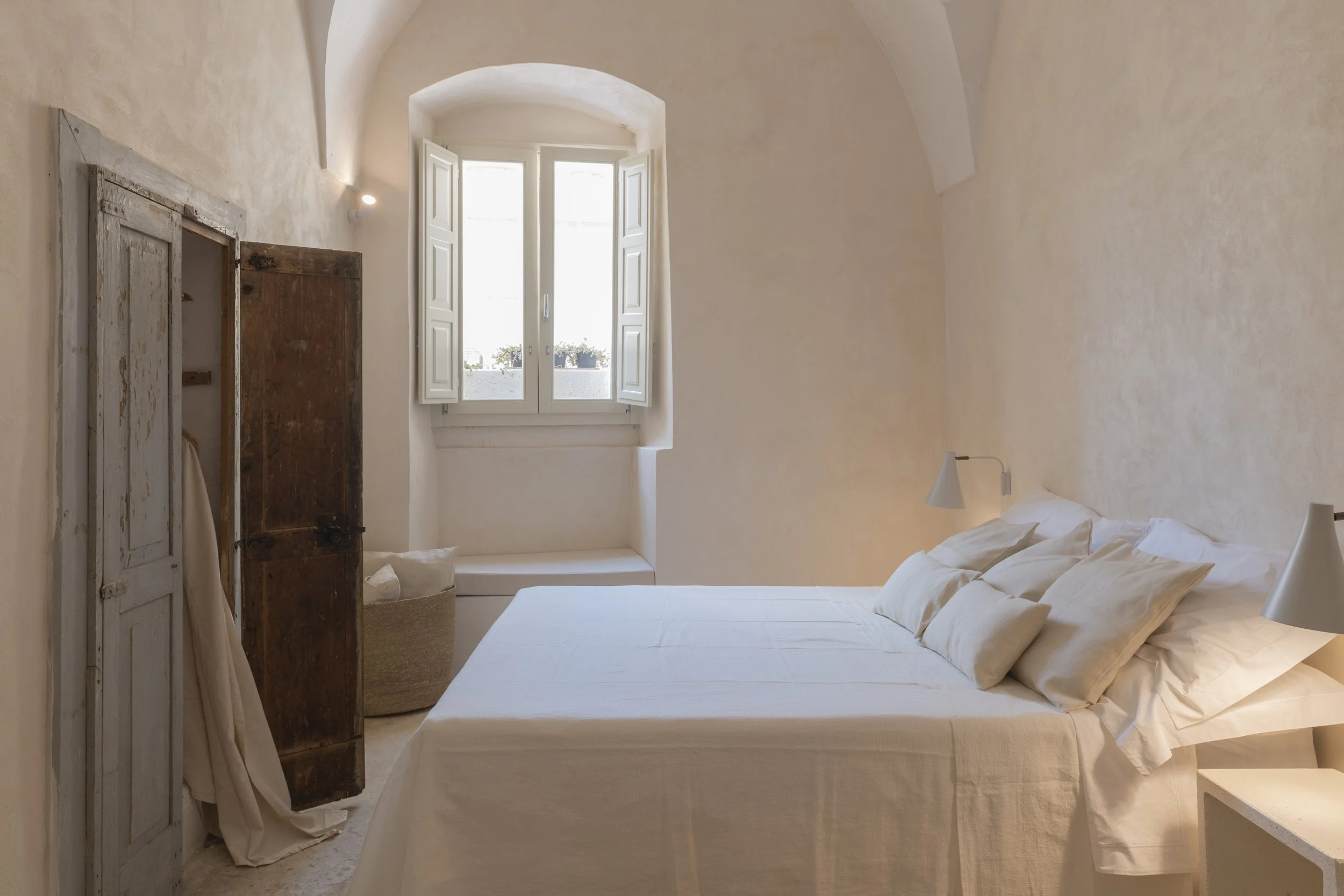 A cozy bedroom with a white bed, beige pillows, and two wall-mounted lamps. There is a window with white shutters allowing natural light, and an antique wooden wardrobe with a partly opened door. The room has soft beige walls and a simple, minimalist
