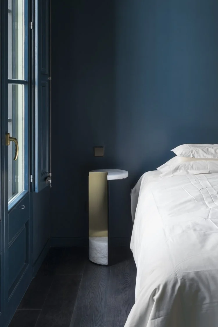 A bedside corner in a bedroom with a dark teal wall, a small round side table, and part of a white bed with white linens.
