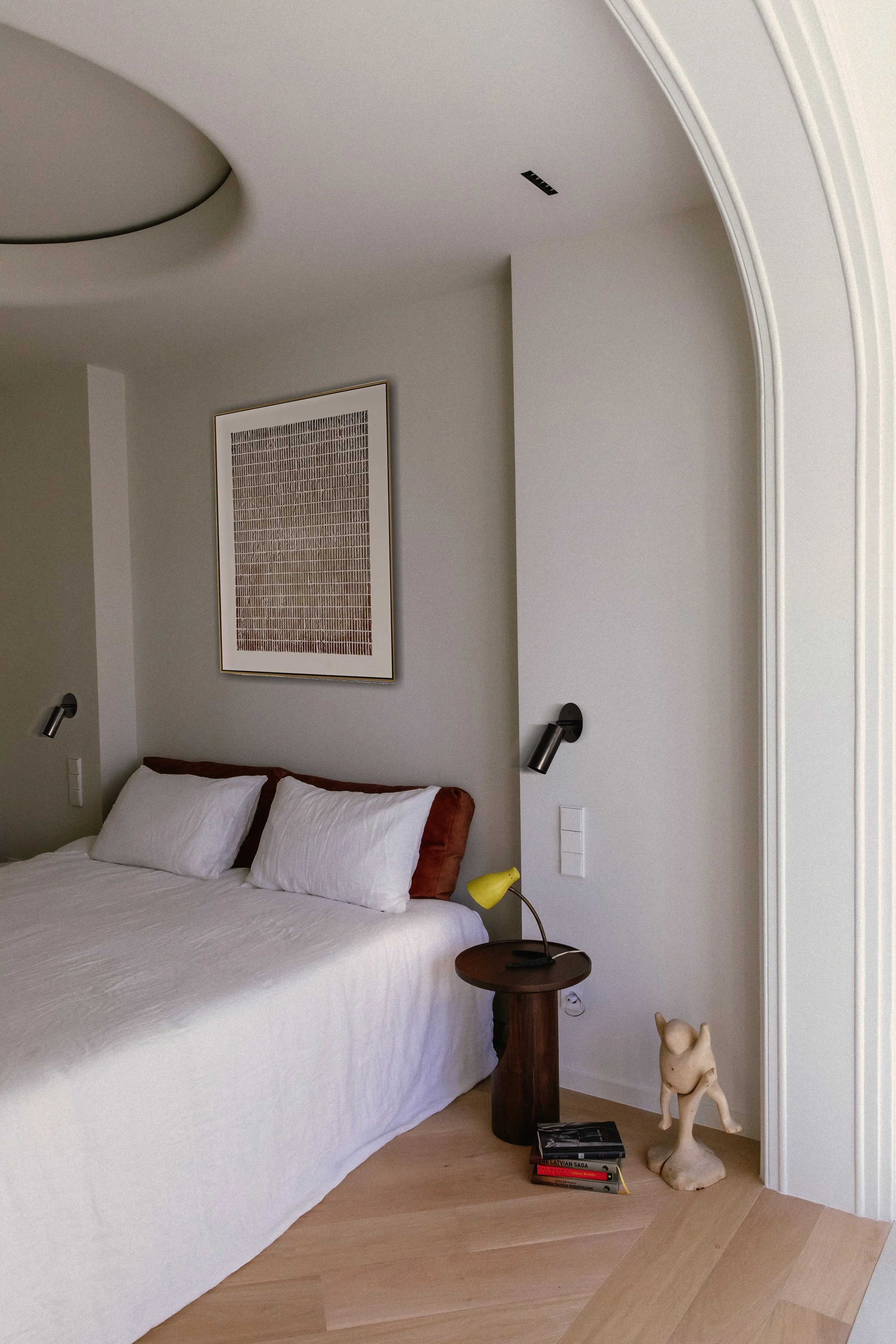 A bedroom with a white bed, two white pillows, and a wooden headboard. There is a round wooden side table with a yellow desk lamp and a sculpture of a cat next to some books on the floor. The walls are painted light, and there are two black reading l