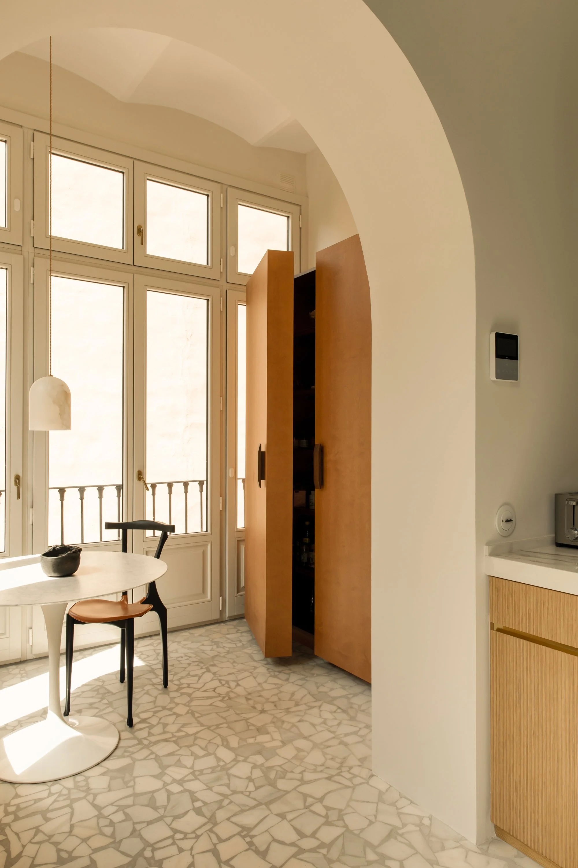 Interior view of a bright kitchen area with large windows, a small round table, a black and brown chair, a wooden cabinet, and a terrazzo floor.