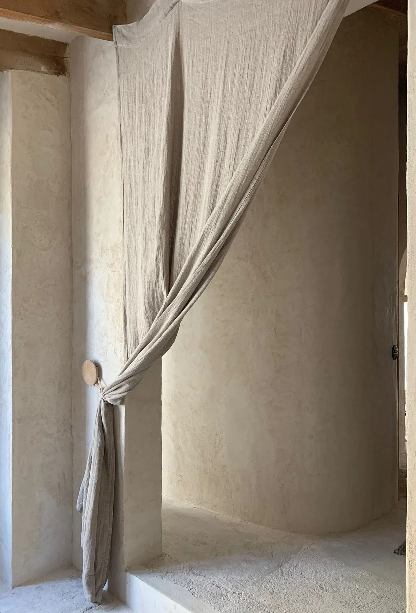 A beige curtain tied back with a round wooden tieback, hanging in front of a curving beige wall and doorway.