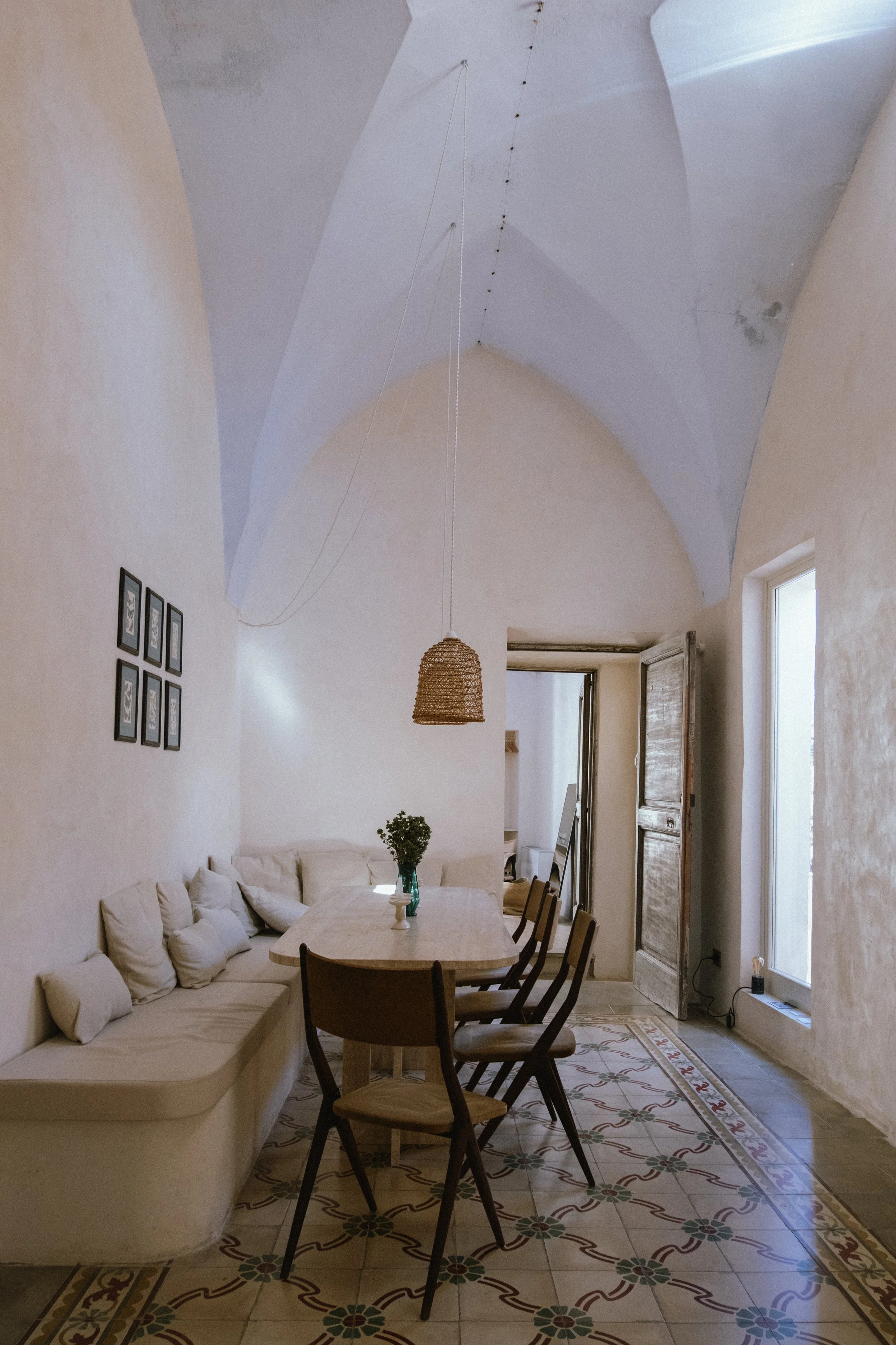 A dining room with a high vaulted ceiling, white textured walls, a large window, a dining table with chairs, and a hanging woven light fixture.