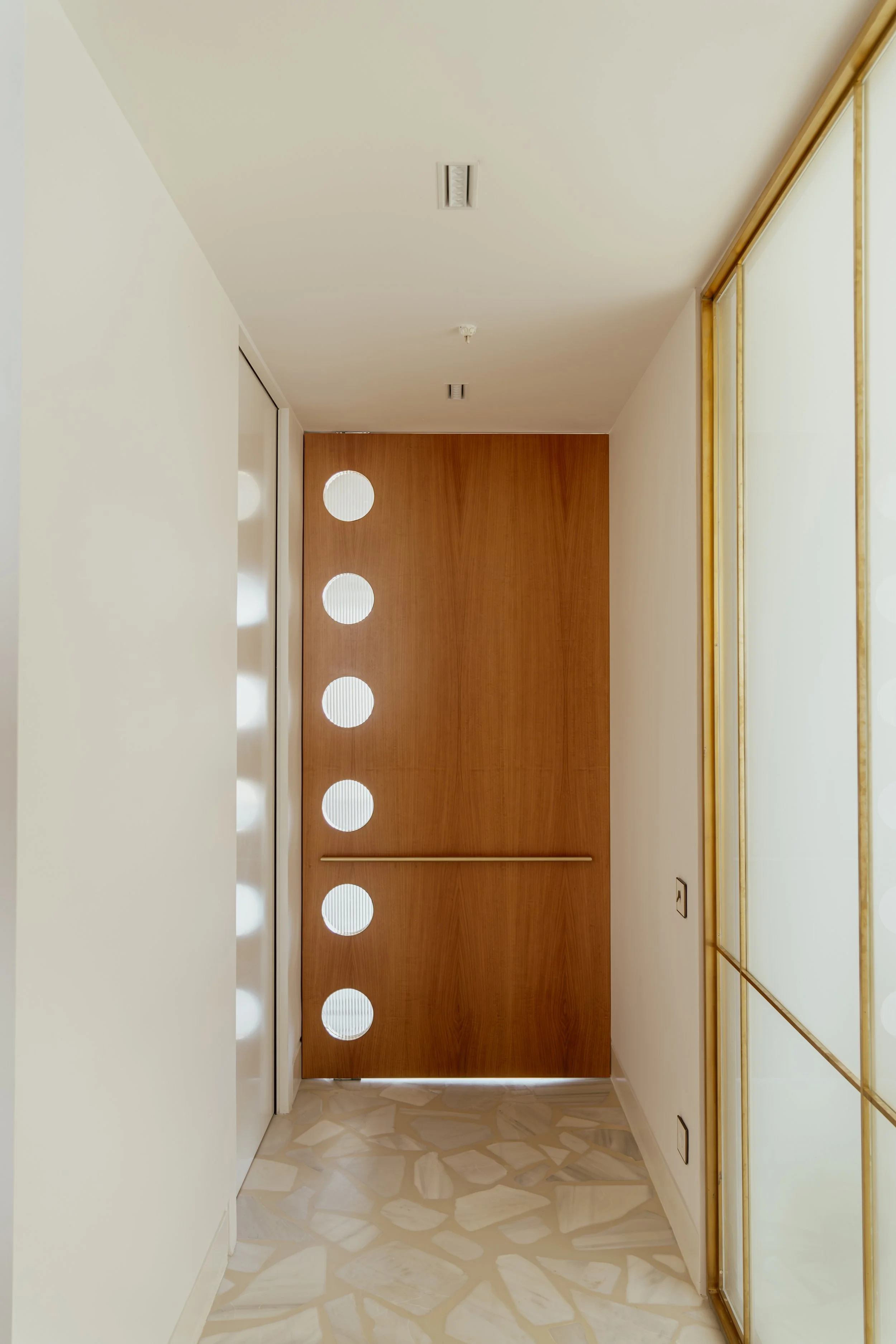 Modern interior hallway with a wooden door featuring circular cutouts, marble floor, and mirrored wall with gold framing.