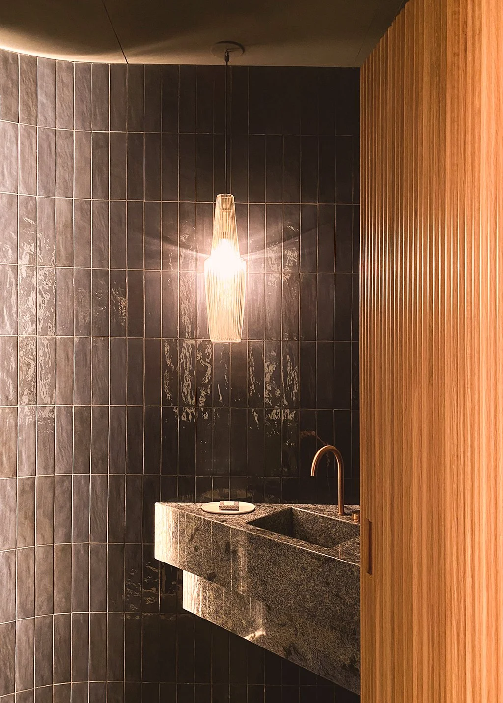Interior view of a modern kitchen or bar area with black tiled wall, a hanging pendant light, a stone sink with a faucet, and a wooden slat divider.