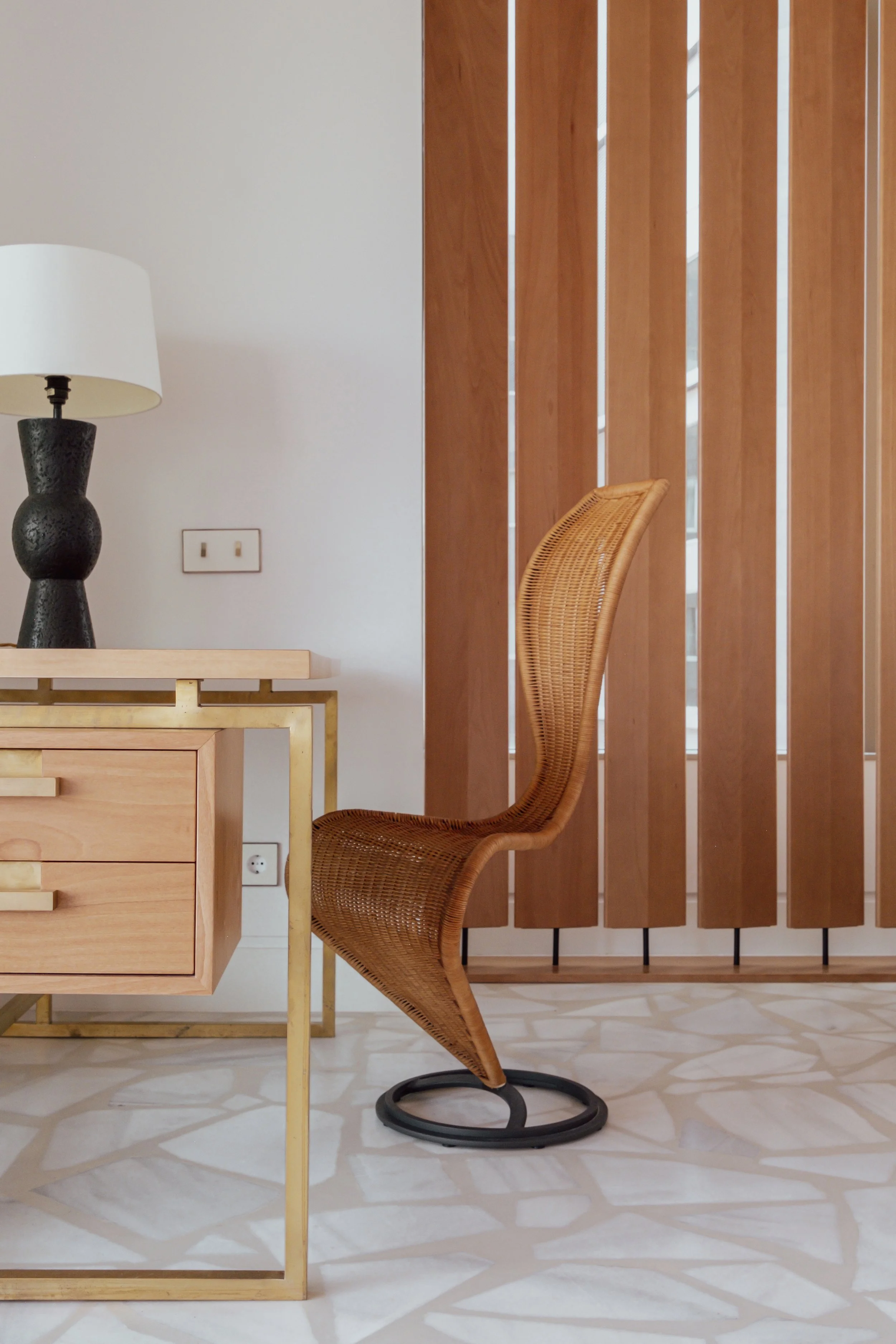 Interior space featuring a wooden desk with gold accents, a black table lamp, a rattan office chair, a wooden panel wall, and tiled flooring.