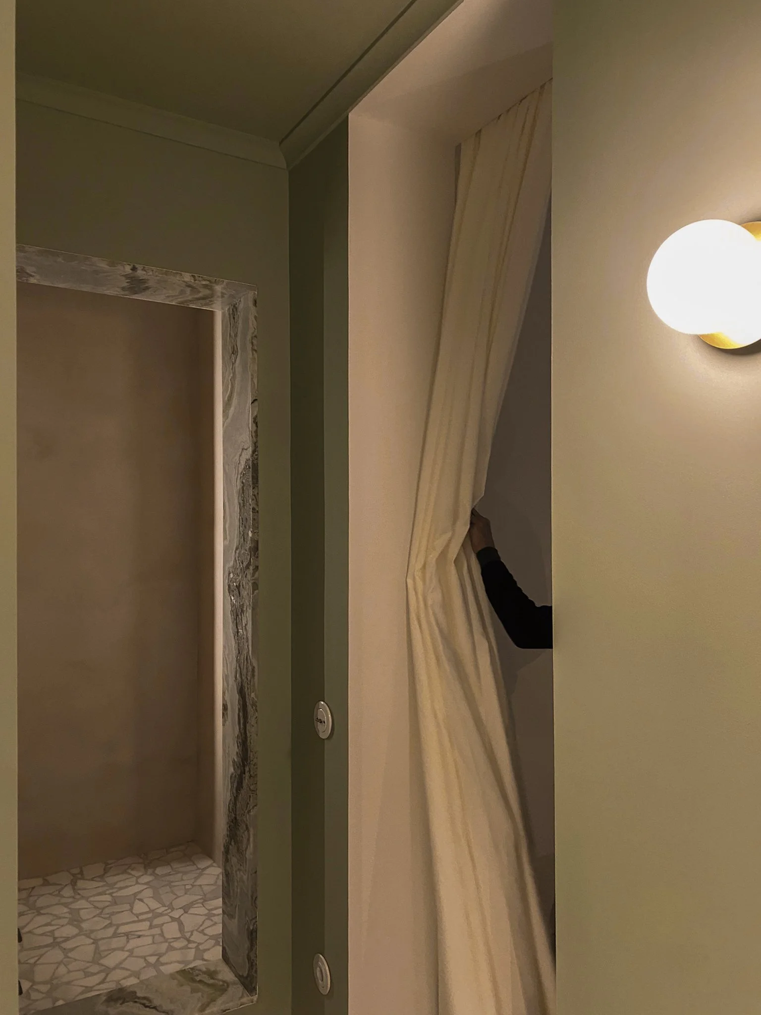 Person partially visible behind a beige curtain in a narrow space, with a wall-mounted round light fixture on the right, and a doorway with marble trim on the left.
