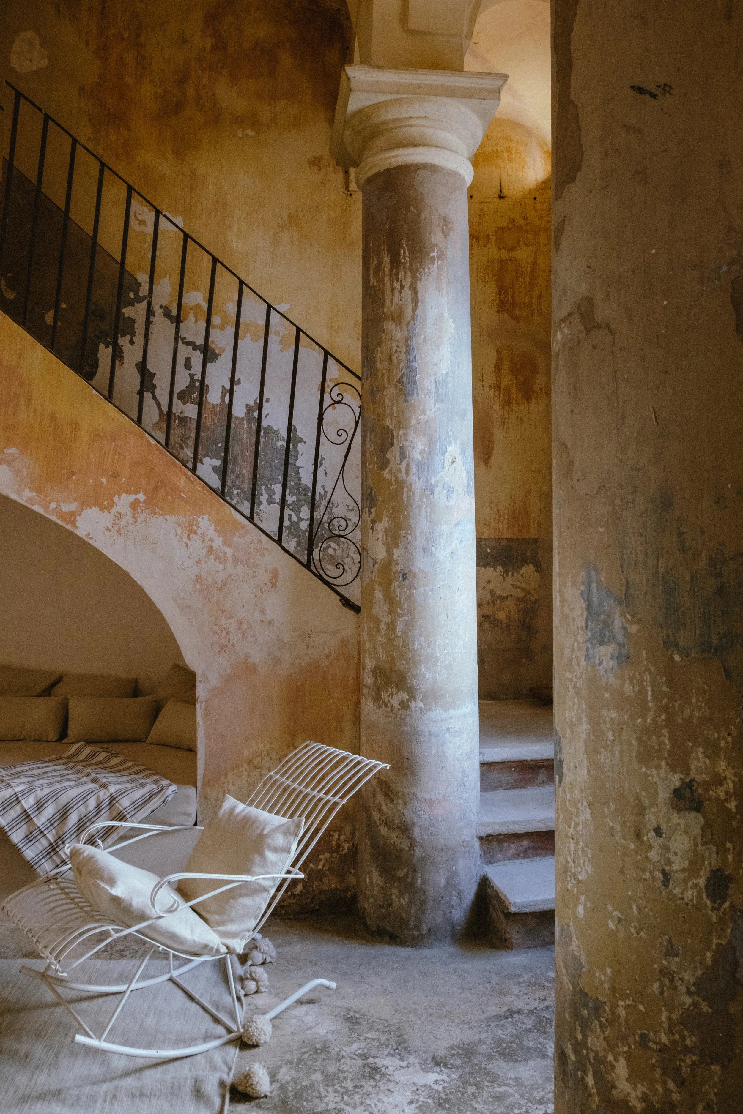 Interior of a rustic, weathered space with peeling yellow paint, a stone spiral staircase, and a white metal rocking chair with pillows and a blanket.