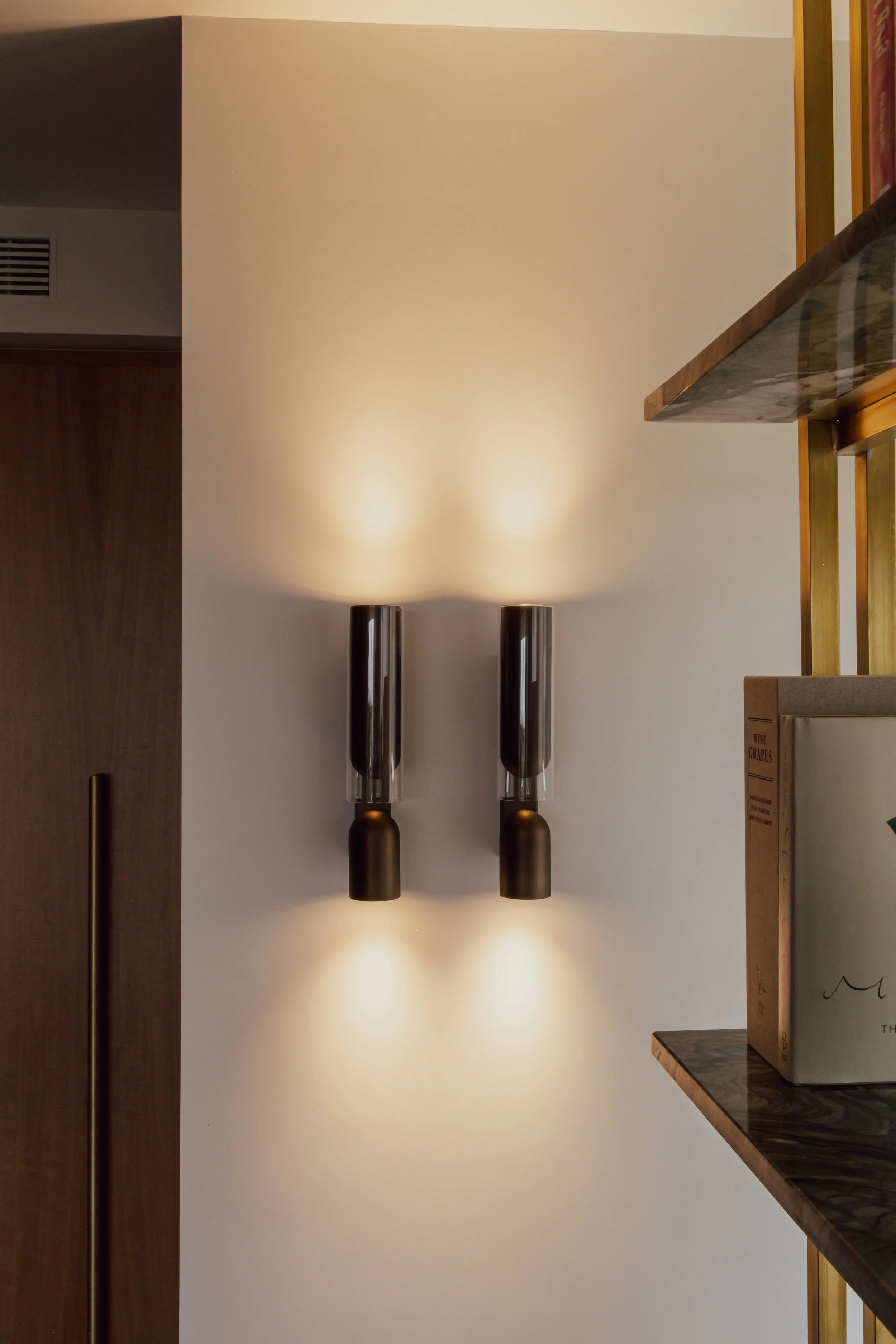 Two modern wall-mounted light fixtures with cylindrical black and transparent glass shades on a cream-colored wall, casting warm light.