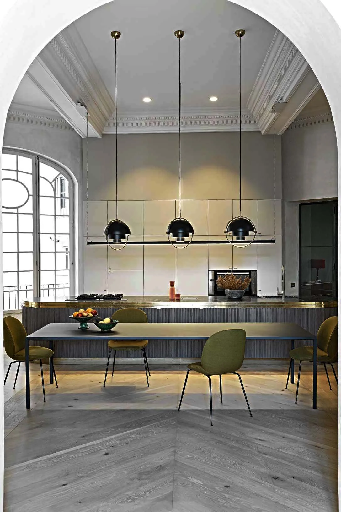 Modern kitchen with high ceilings, large arched window, three black pendant lights, white tiled wall, dark wood cabinets, black dining table, green chairs, and a wooden floor.