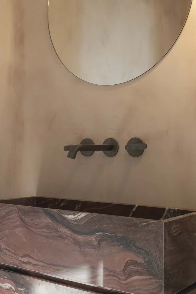 A bathroom corner with a marble countertop and a beige wall. A modern black faucet and a round wall mirror are visible.