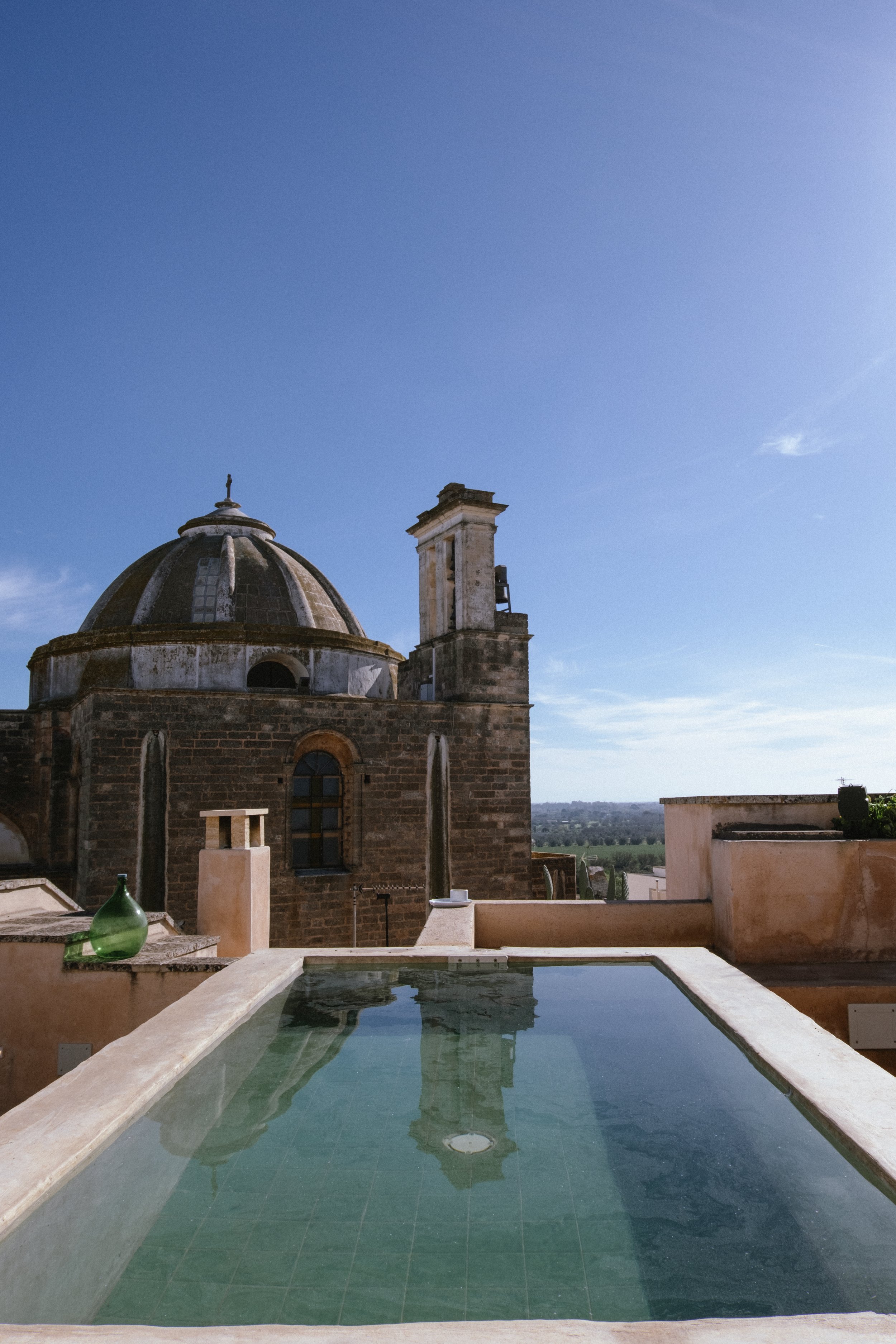 Rooftop view of an old church with a domed roof, a tower, and surrounding landscape under a clear blue sky, with a reflection in a small pool of water in the foreground.