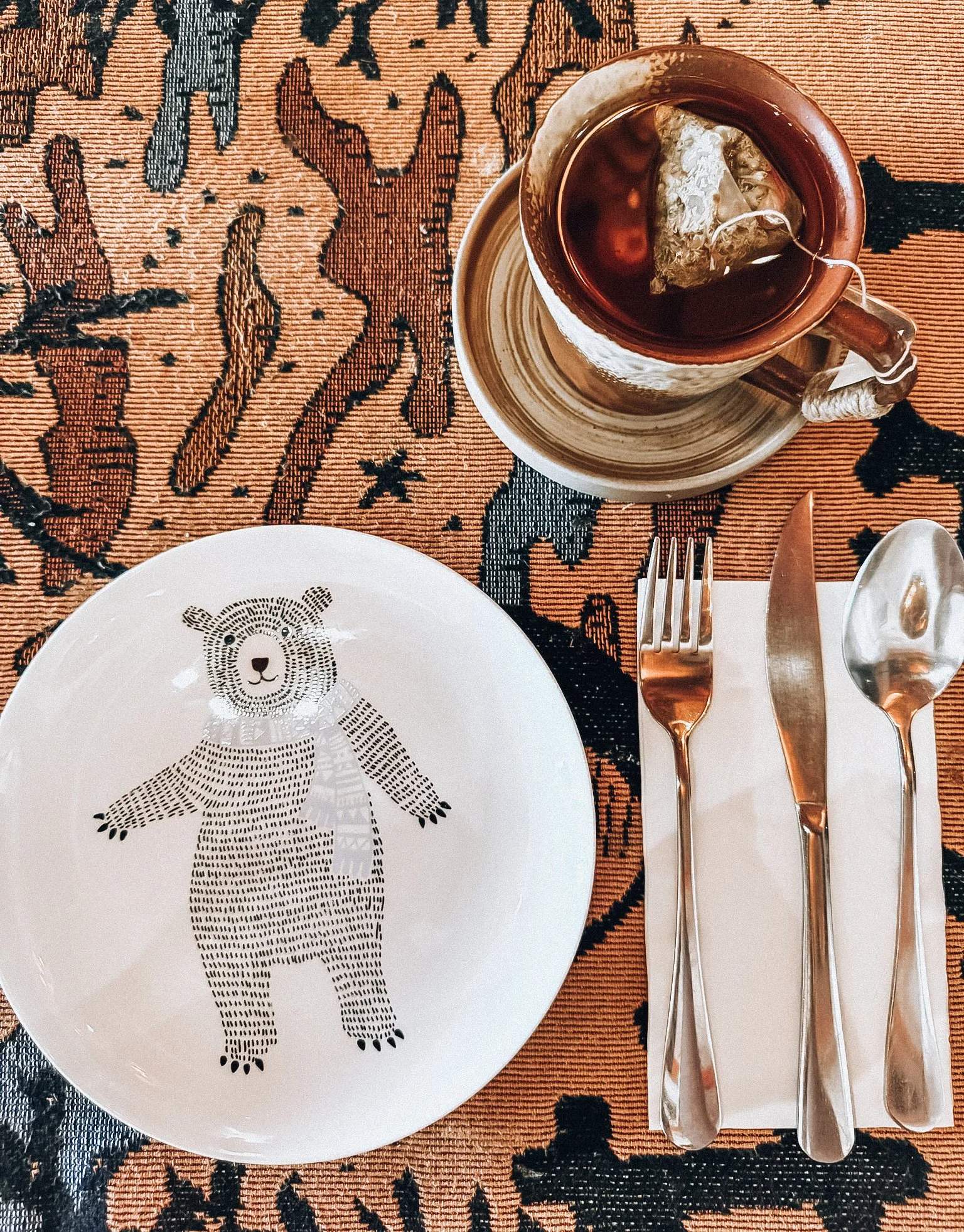 A white plate with a black bear illustration, a set of silverware on a white napkin, and a cup of tea on a patterned tablecloth.