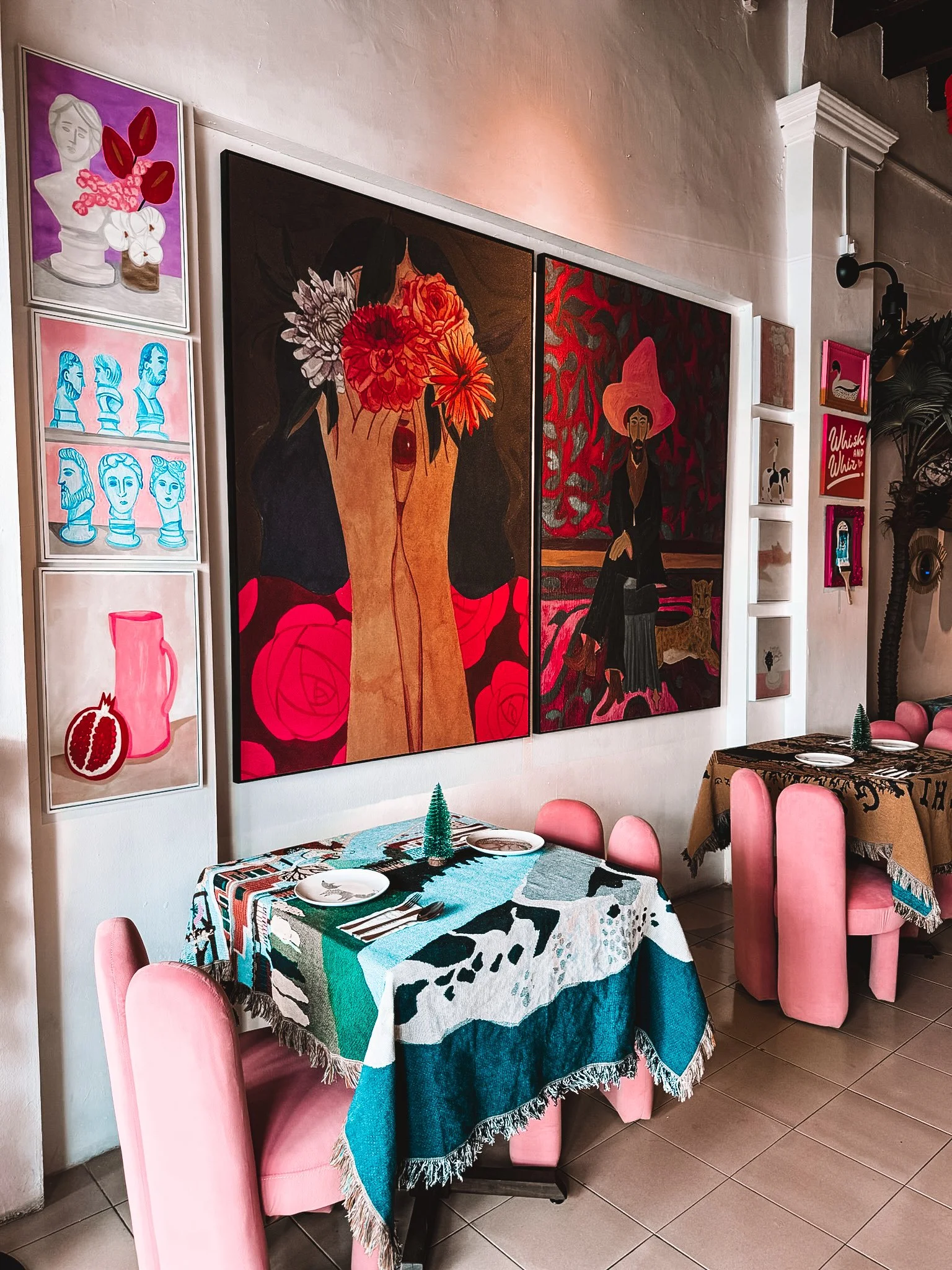 A cozy restaurant interior with pink chairs, colorful patterned tables, and wall art featuring bright paintings of women, flowers, and abstract figures.