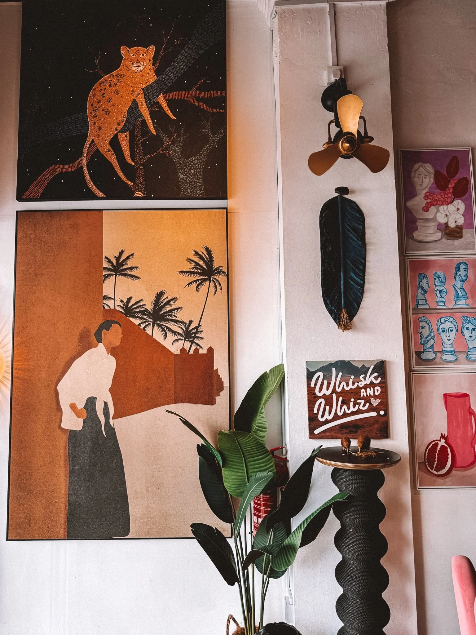 Interior wall with various art pieces, including a painting of a leopard on a tree, a painting of a person with palm trees and a castle silhouette, a sign that reads 'Whisk and Whiz', a feather wall hanging, and some framed illustrations of faces, flowers, and a pomegranate, along with a stylish black textured pedestal with a plant, a ceiling fan, and a beige concrete wall.