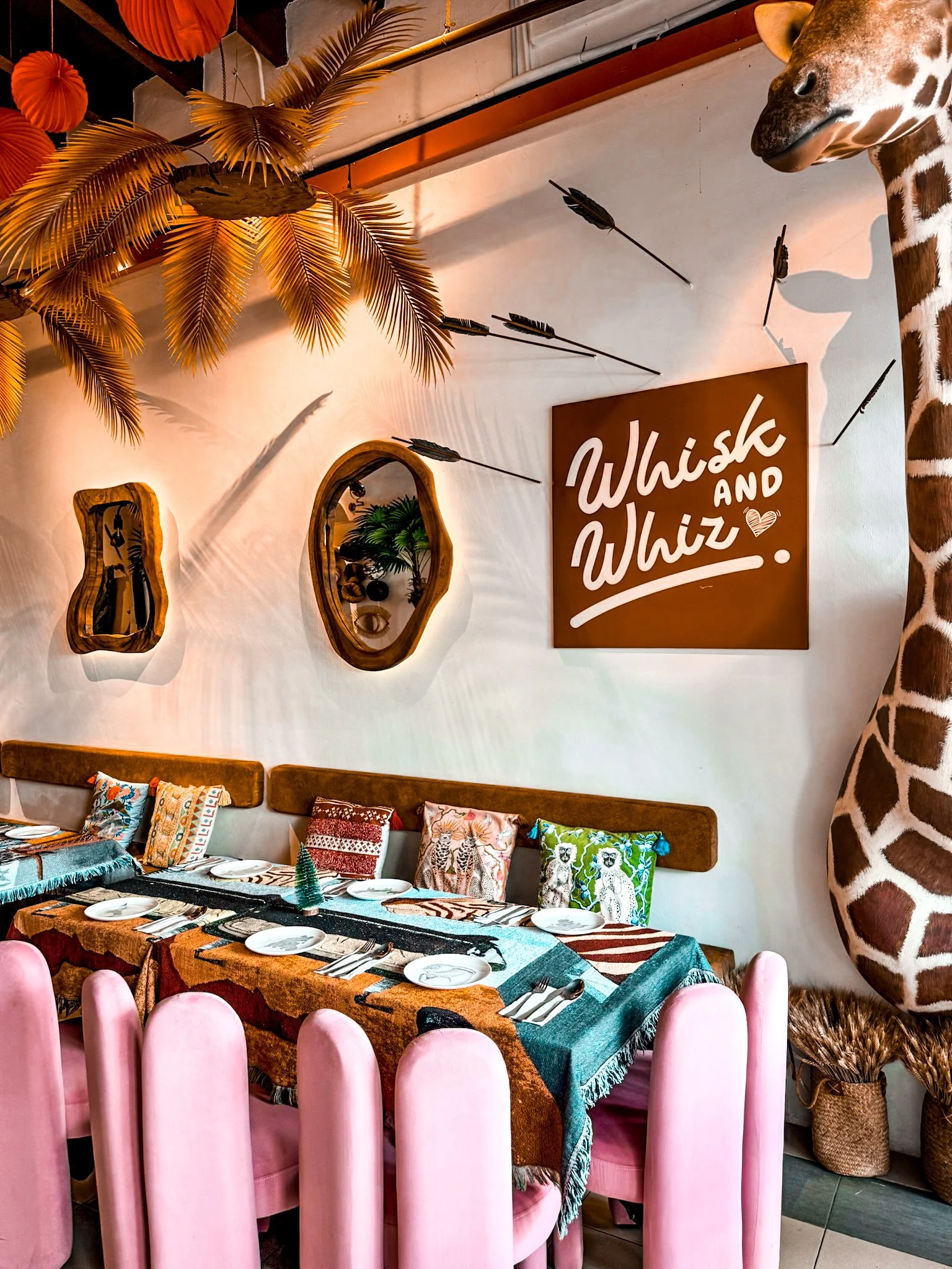 A jungle-themed restaurant interior with tropical decor, a giraffe sculpture on the right, a sign that says 'Whisk and Whiz', and a long dining table set with plates, cutlery, pink chairs, and colorful cushions on a bench.