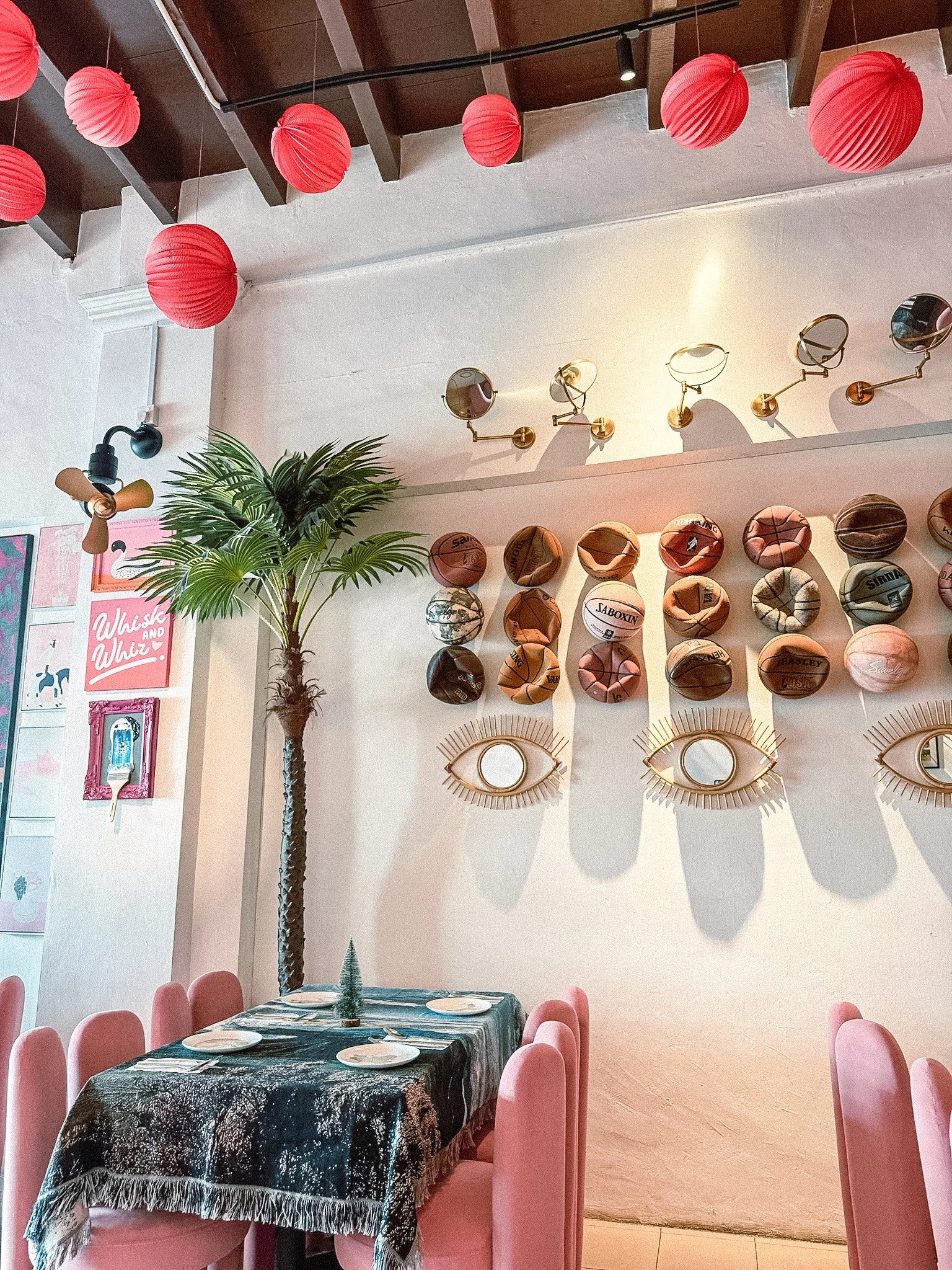 Decorated dining area with pink chairs, a table with a dark tablecloth, a small Christmas tree centerpiece, pink paper lantern ceiling lights, wall-mounted spherical mirrors, and a wall display of basketballs arranged in a pattern with decorative eye-shaped mirrors.