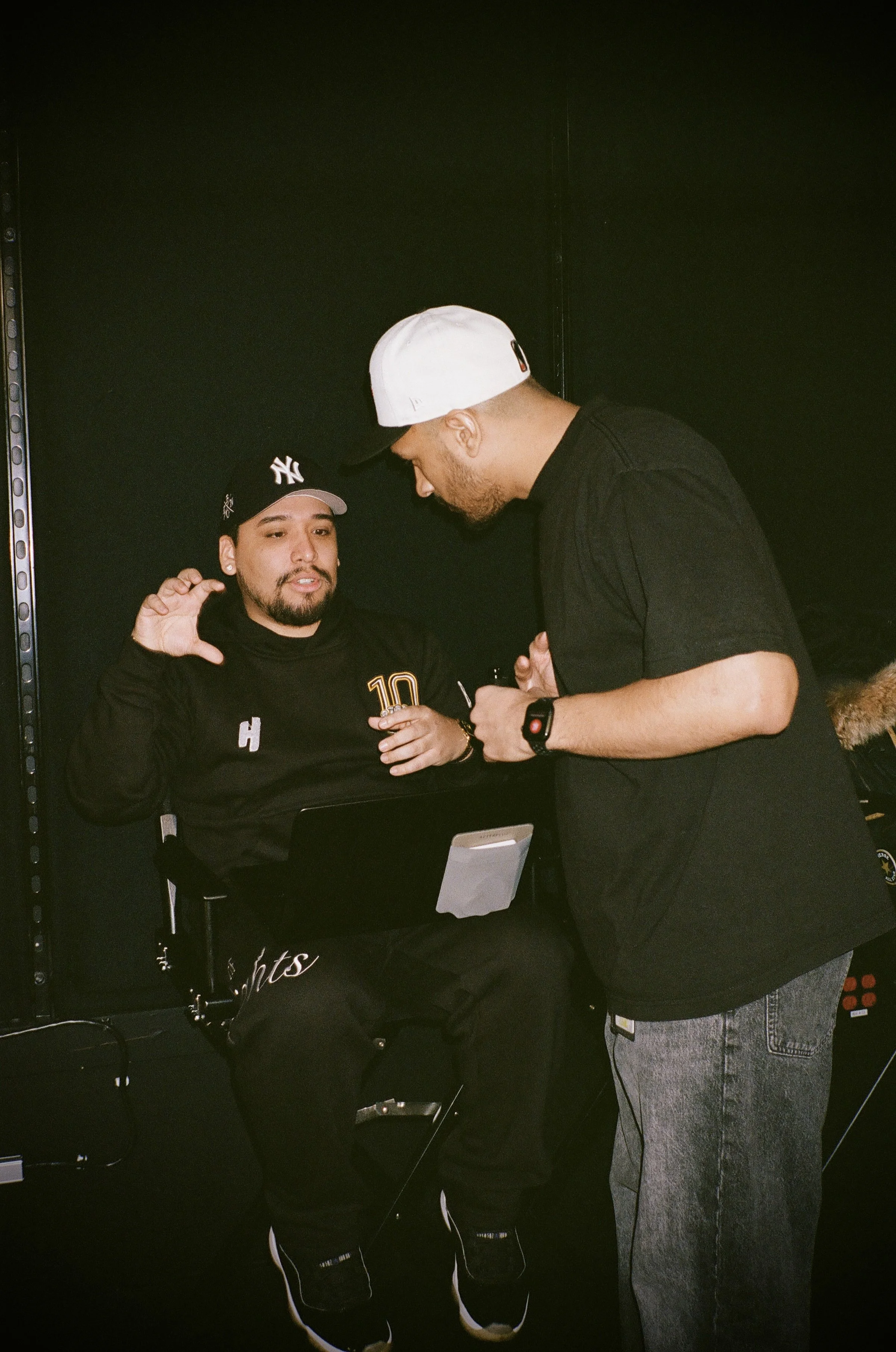 Two men are having a conversation in a dark room, one seated and one standing, with black walls around them.