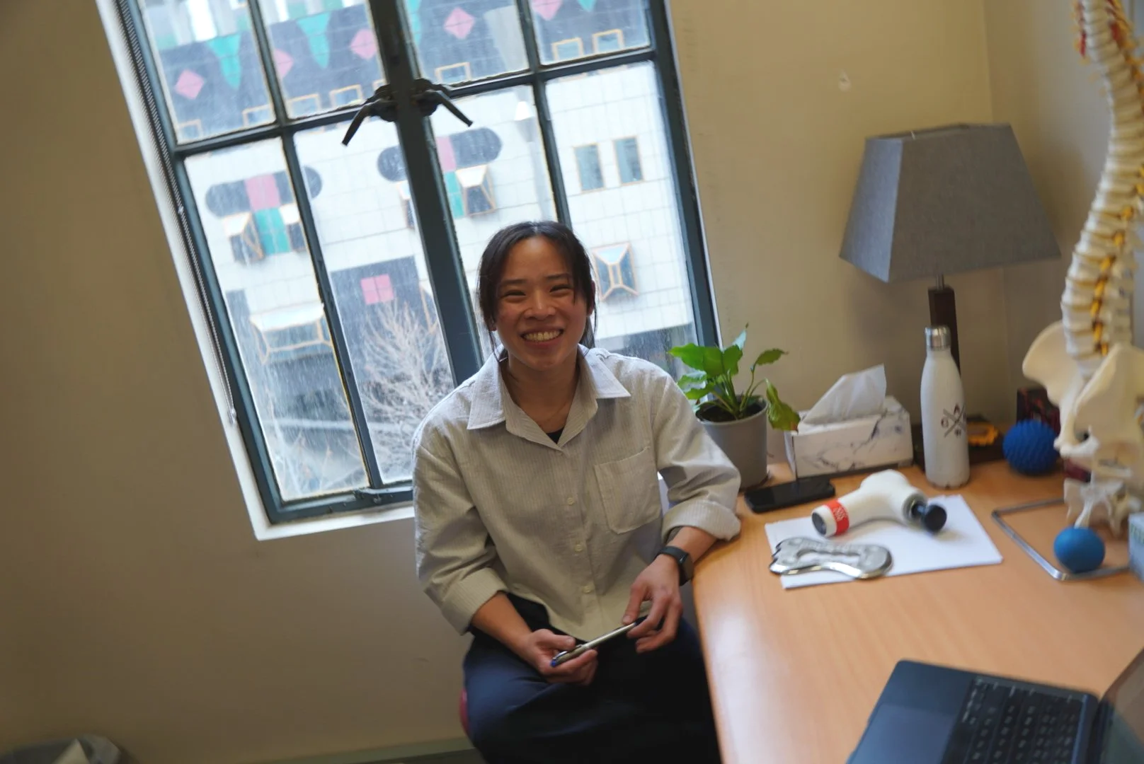 A woman sitting at a desk, smiling, with a window behind her and various objects on the desk including a potted plant, a white water bottle, and small items.