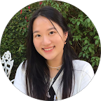 A smiling young woman with long dark hair, outdoors surrounded by green foliage and white flowers, in a circular frame.