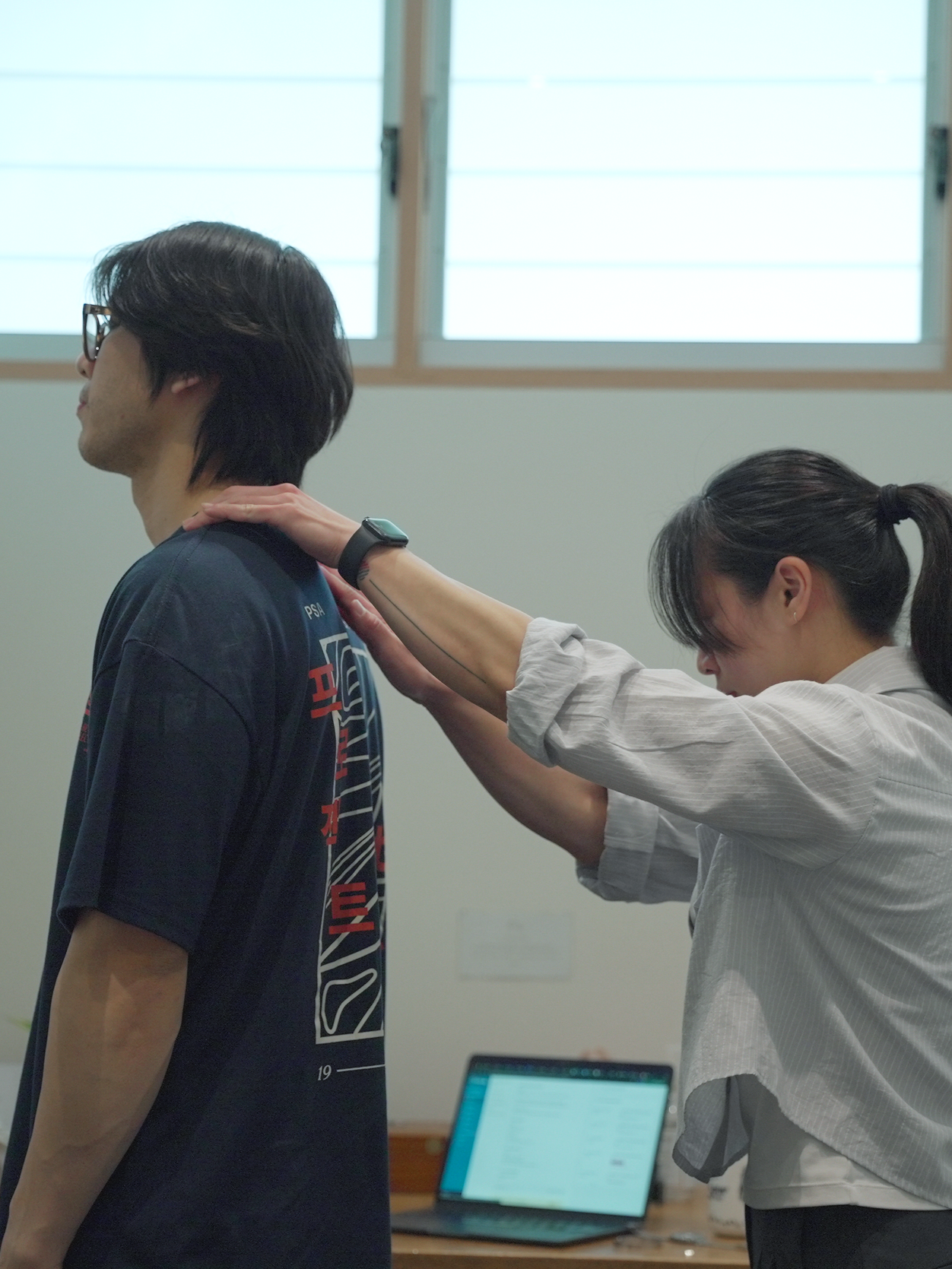 A woman is performing a neck stretch on a young man with glasses and dark hair, who is standing upright in a room with windows and a laptop on a table in the background.